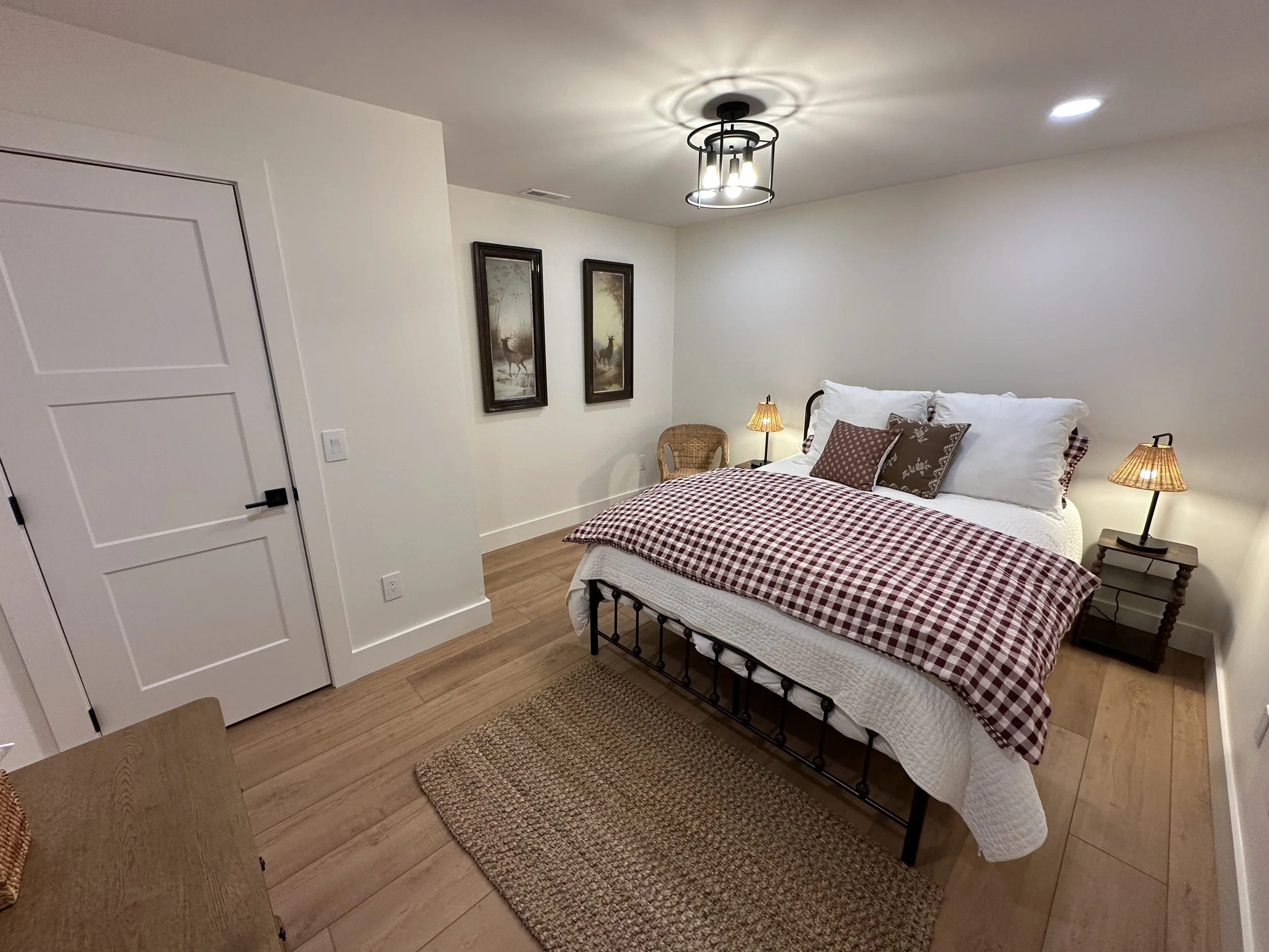 A cozy bedroom with a neatly made bed featuring a red and white checkered comforter, surrounded by wooden side tables with small lamps, two framed nature prints on the wall, and a wicker chair in the corner. Hardwood floors and a simple ceiling light