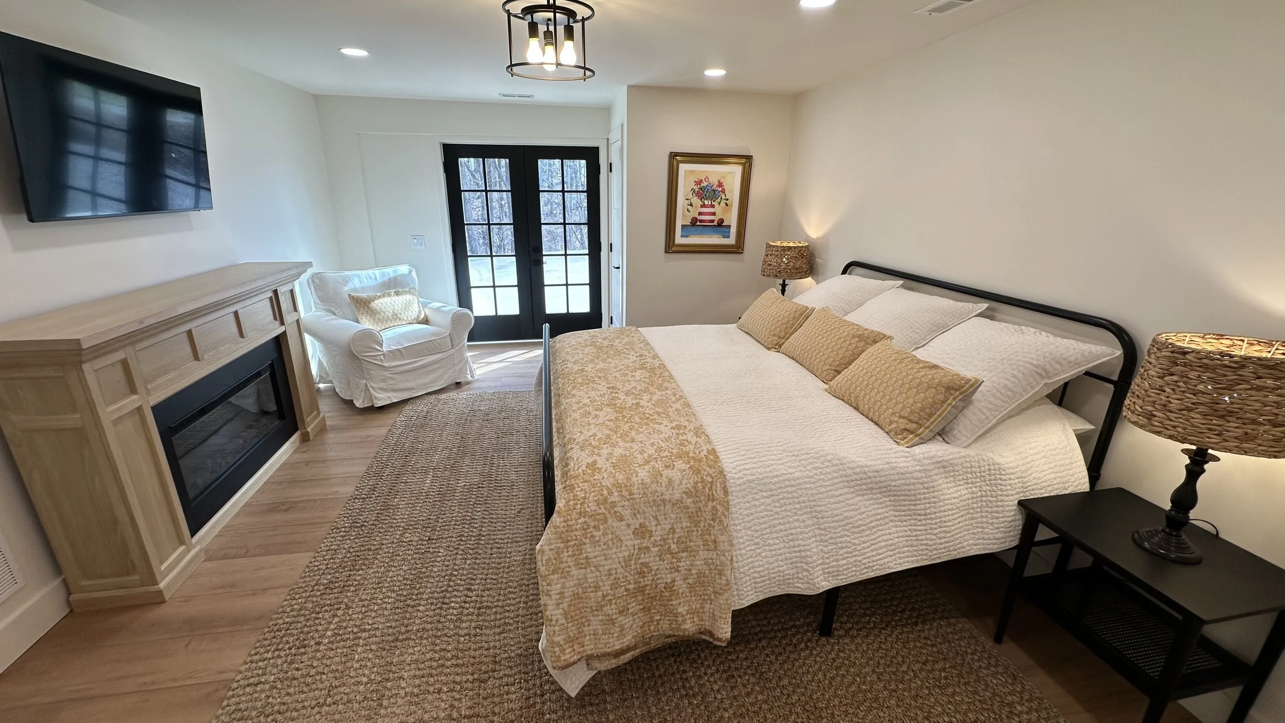 A bedroom with a queen-sized bed, beige pillows, and a beige bedspread. A black nightstand with a woven lamp, a white armchair near black double glass doors, and a wall-mounted flat-screen TV above a light wooden fireplace mantle. There's a framed pi
