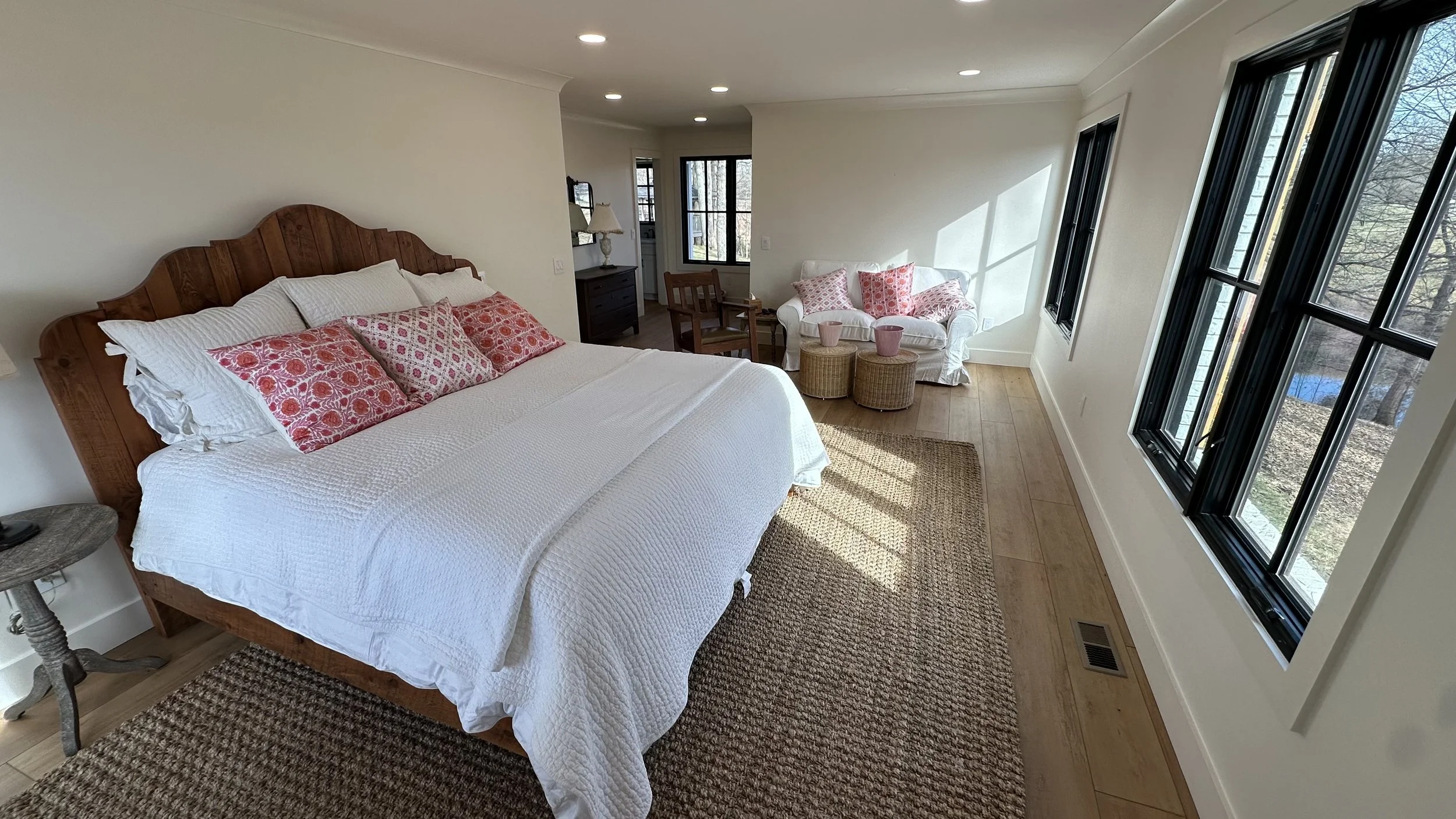 A bedroom with a wooden bed frame, white bedding, and pink patterned pillows. There is a small side table with a lamp, a sitting area with a white sofa with pink pillows, two wicker stools, and large windows with black frames letting in natural light