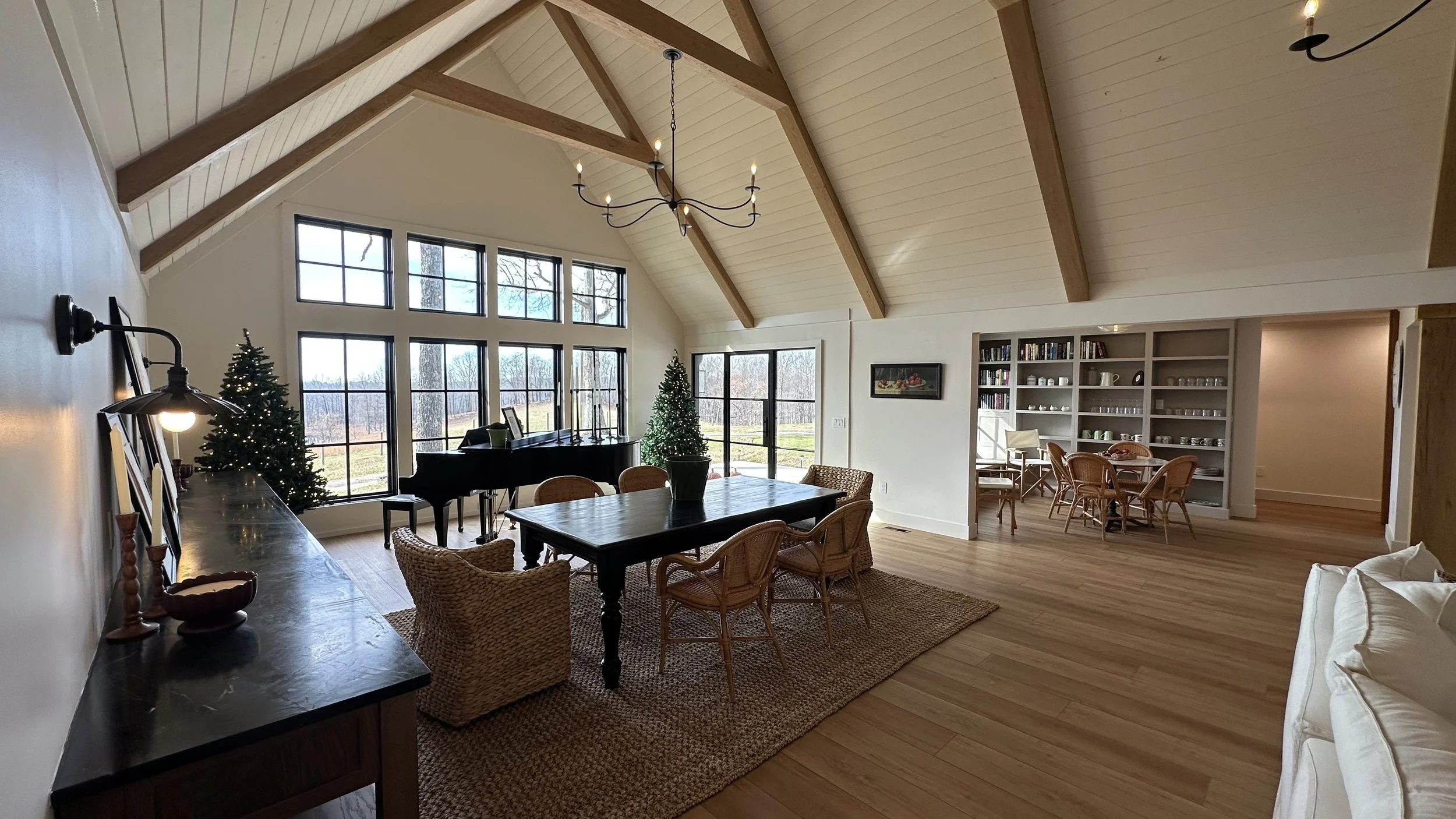 Interior of a spacious living and dining area with high vaulted ceilings, large windows, a black grand piano, decorated Christmas trees, a dining table with wicker chairs, and built-in shelves filled with dishes and glassware.