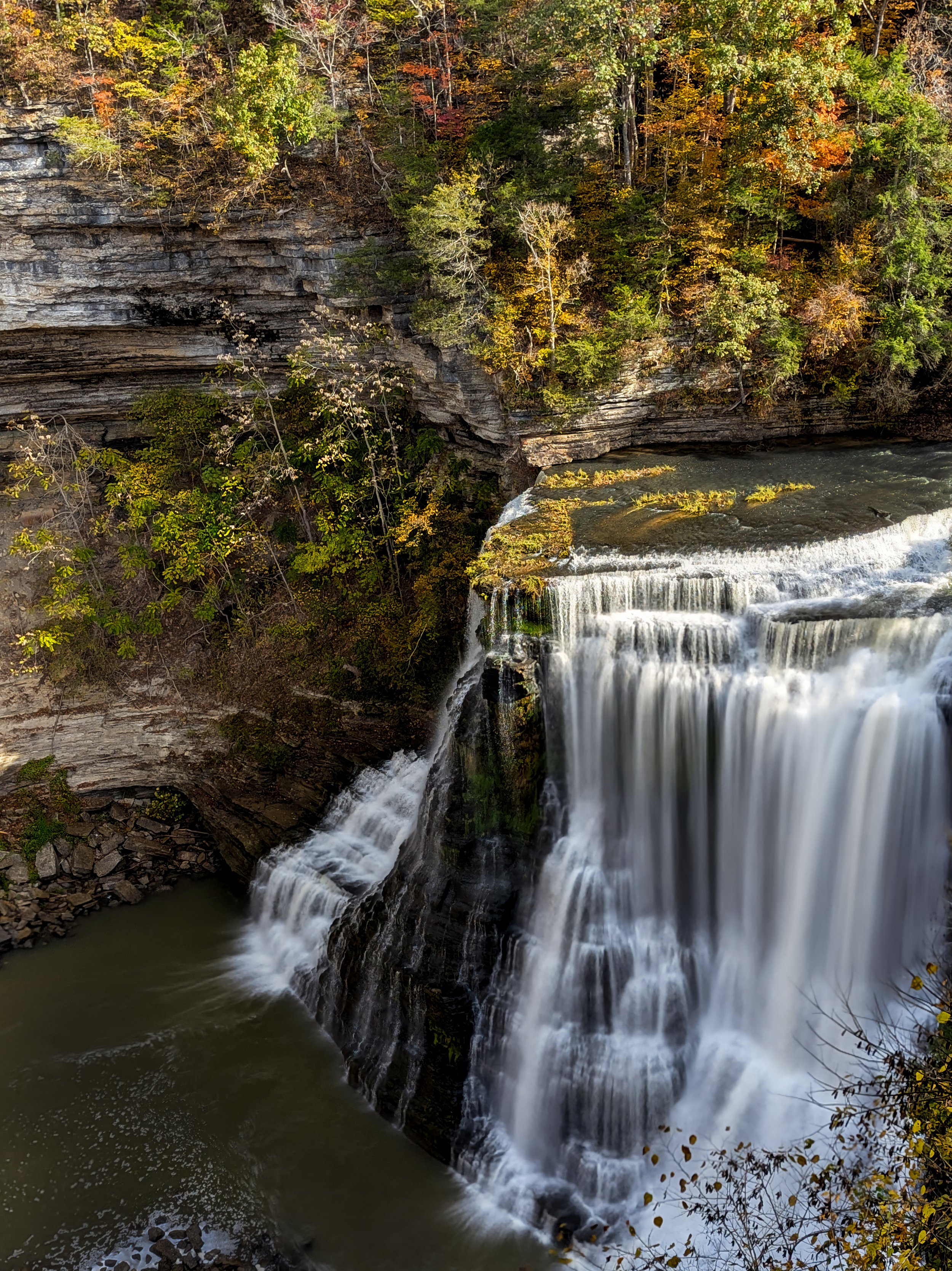 A waterfall cascading over rocks into a river, surrounded by autumn trees with colorful foliage.