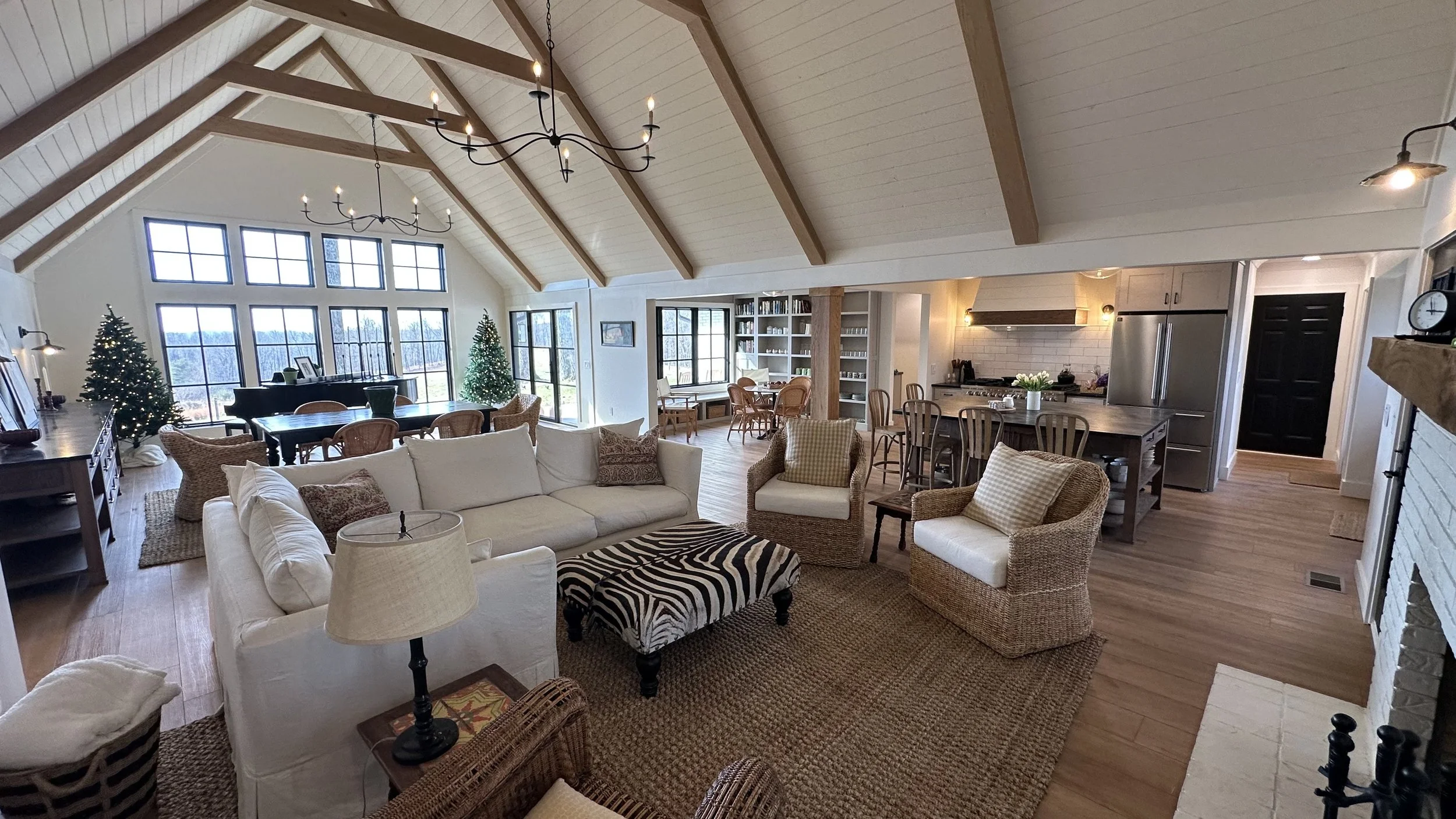 Open-concept living and dining area with vaulted ceiling, large windows, and a kitchen, decorated for Christmas with two Christmas trees, a central white sofa, wicker chairs, a zebra-print ottoman, and wooden furniture.