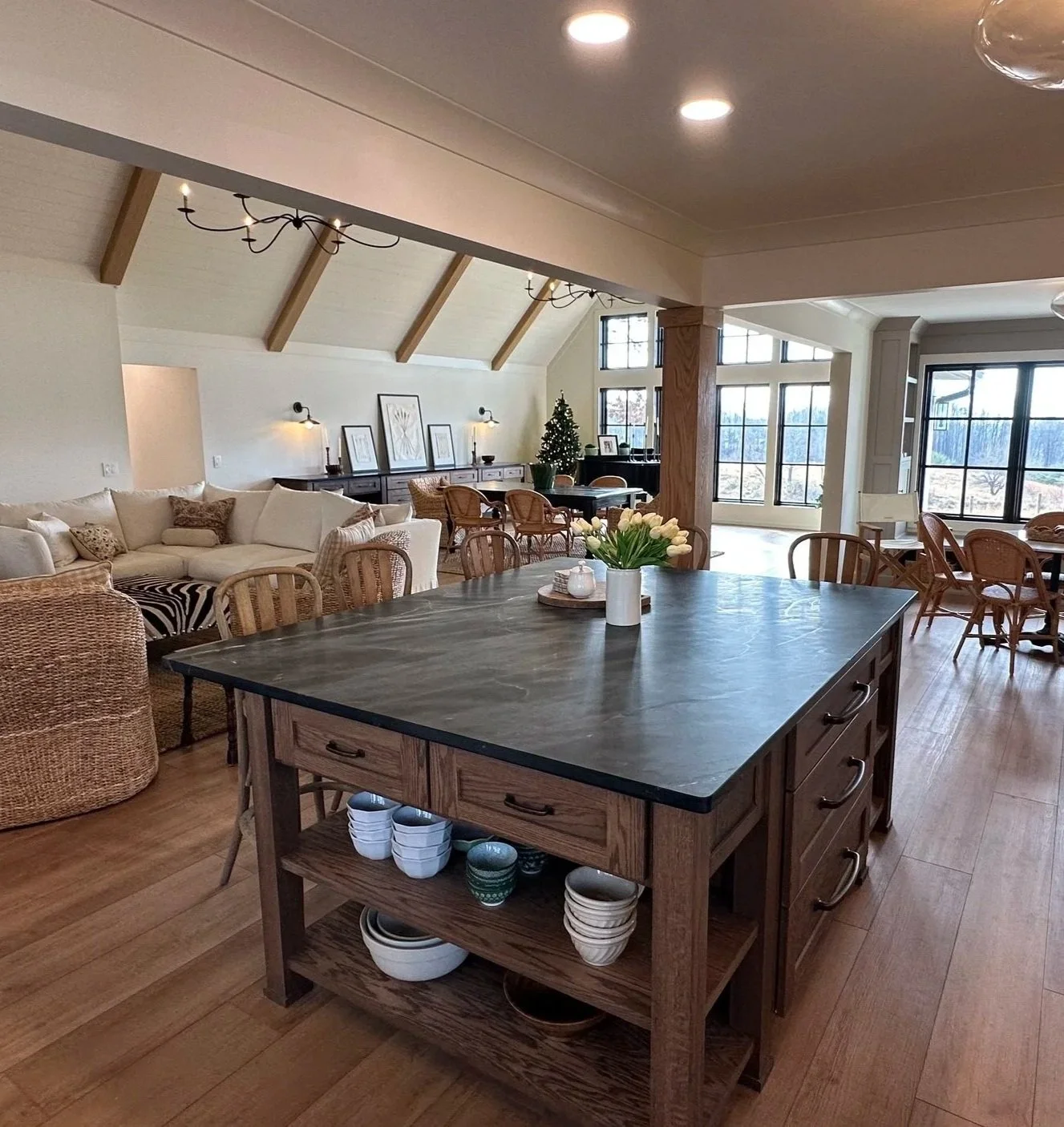 Open living space with a large kitchen island with a dark countertop, surrounded by wooden chairs, with a vase of white tulips on top. In the background, there's a cozy sitting area with a white sectional sofa, wicker armchair, wall-mounted lamps, framed artwork, a Christmas tree, and large windows letting in natural light.