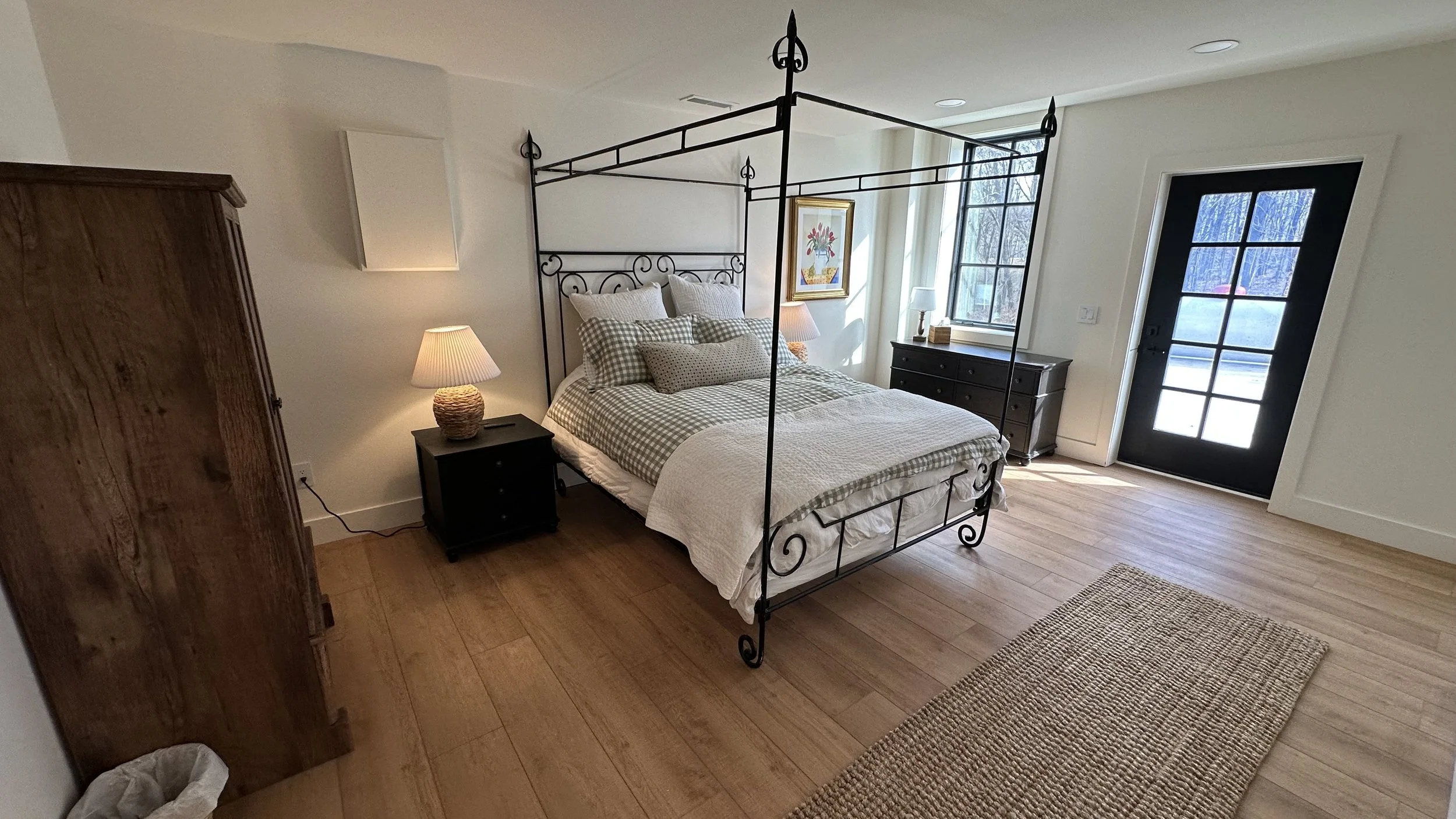 A bedroom with a black metal canopy bed, wooden nightstands with lamps, a dresser by the window, and a rug on wooden floor. A black door with glass panes is at the front.