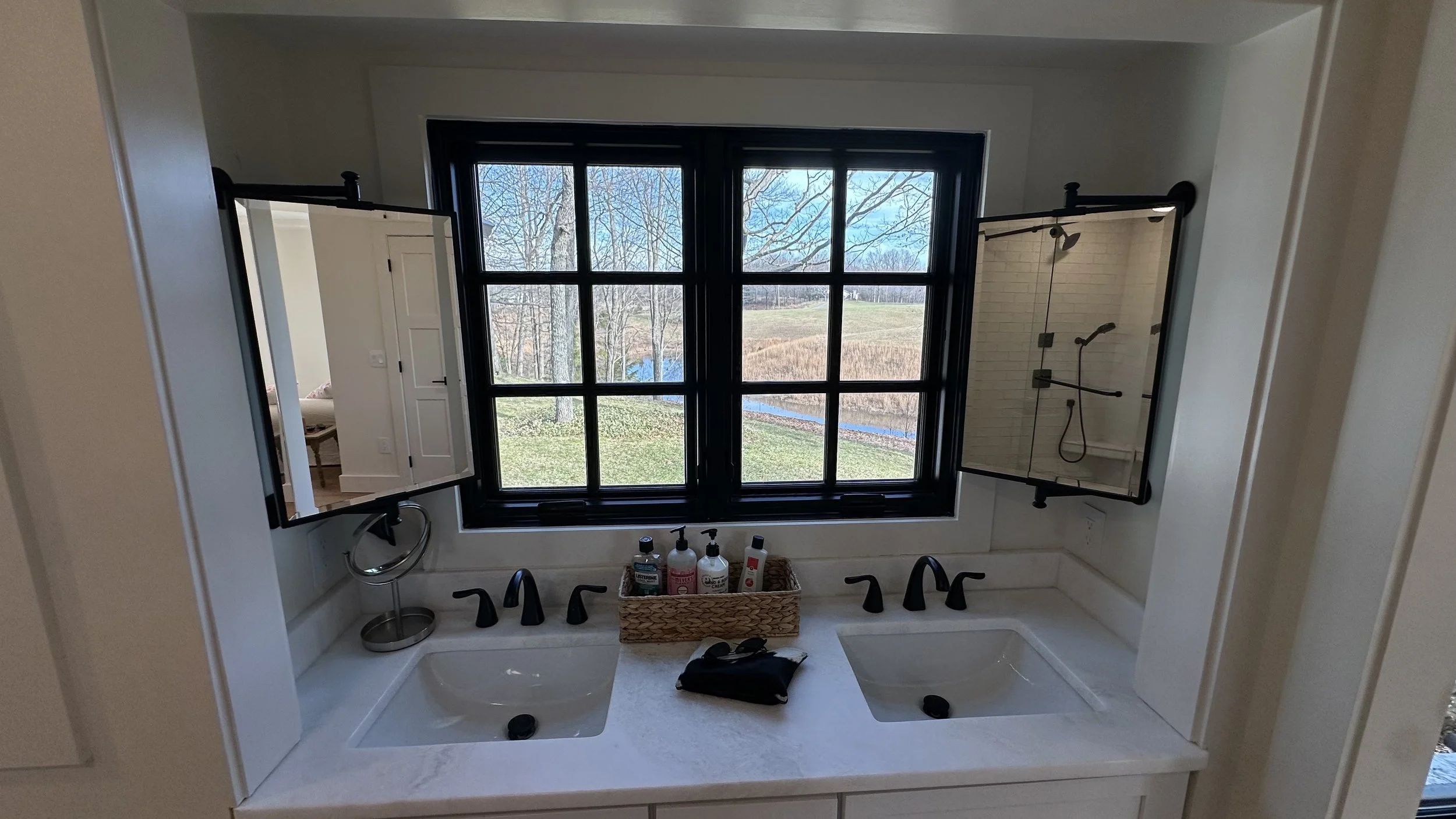 Bathroom vanity with two sinks, two black-finished faucets, a large window overlooking a backyard with trees and grass, and two mirrors, one on each side of the window, reflecting parts of the room.