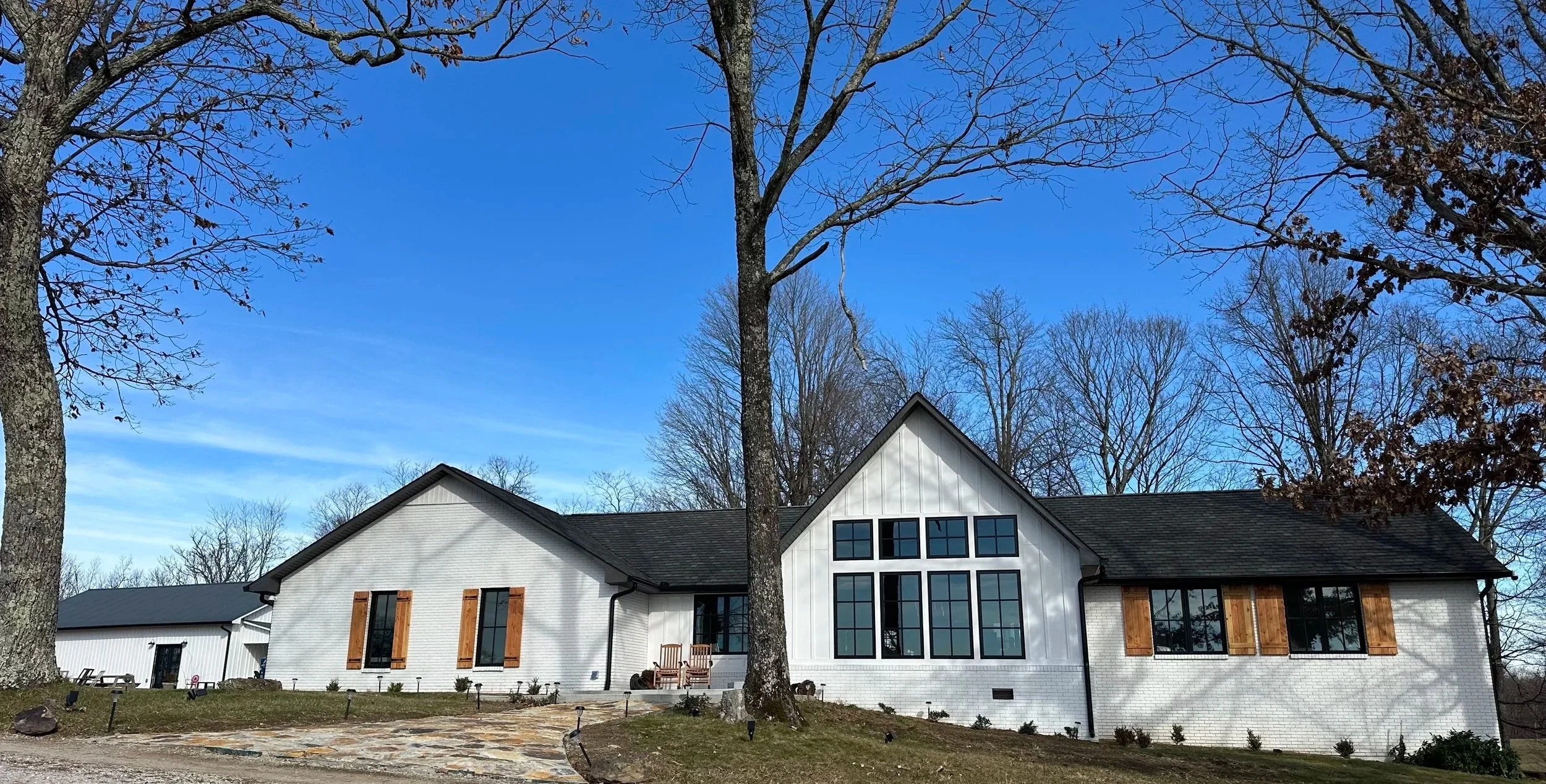 Modern white house with black window frames and wooden shutters, set against a clear blue sky and surrounded by leafless trees.