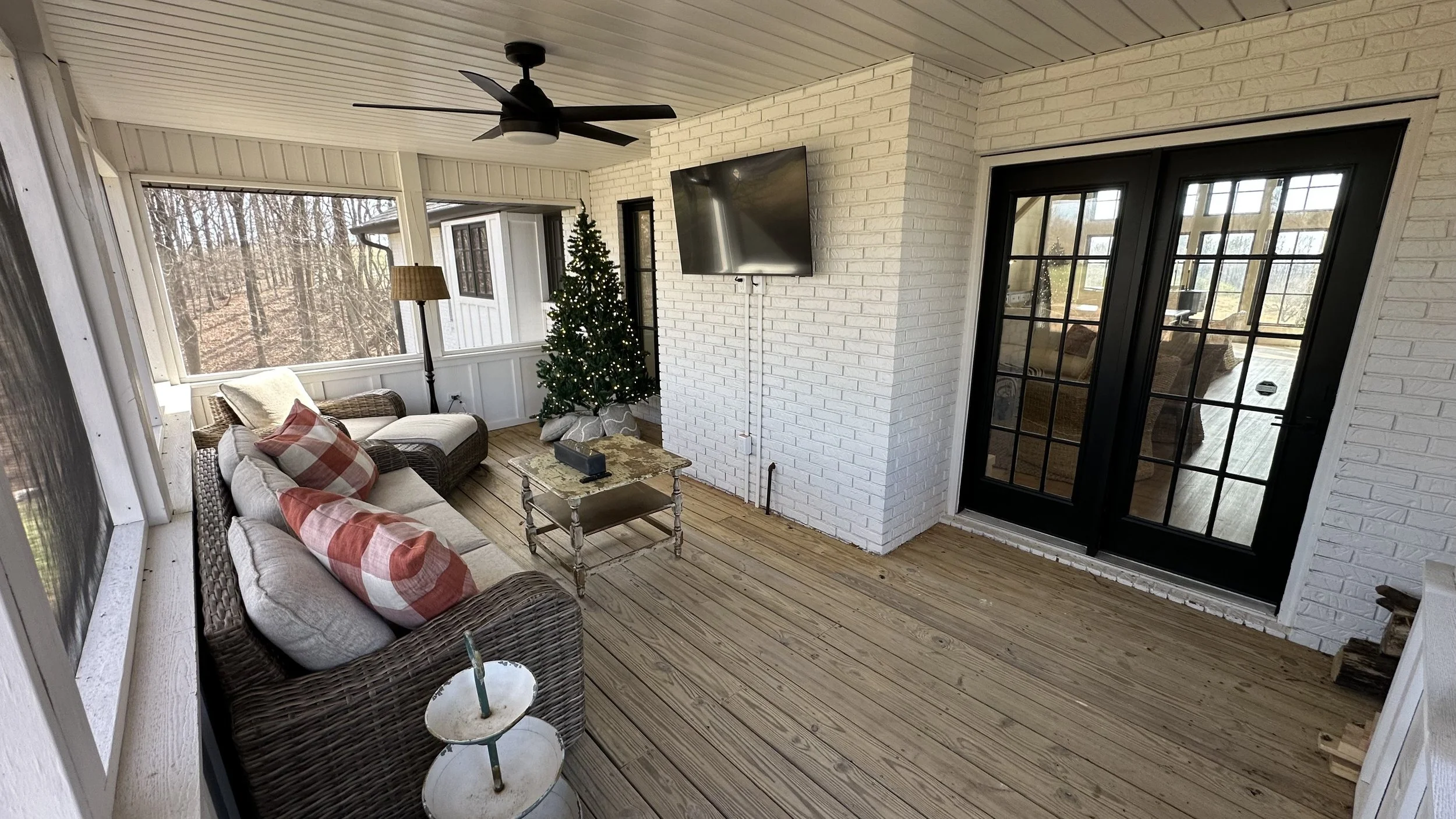 Sunroom with wicker sofa, plaid pillows, small table, Christmas tree, floor lamp, ceiling fan, white brick walls, large windows, black French doors, wooden floor.