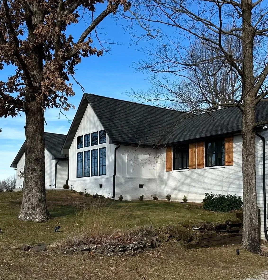 A white house with large windows and wooden shutters, surrounded by leafless trees, on a grassy lawn under a partly cloudy blue sky.