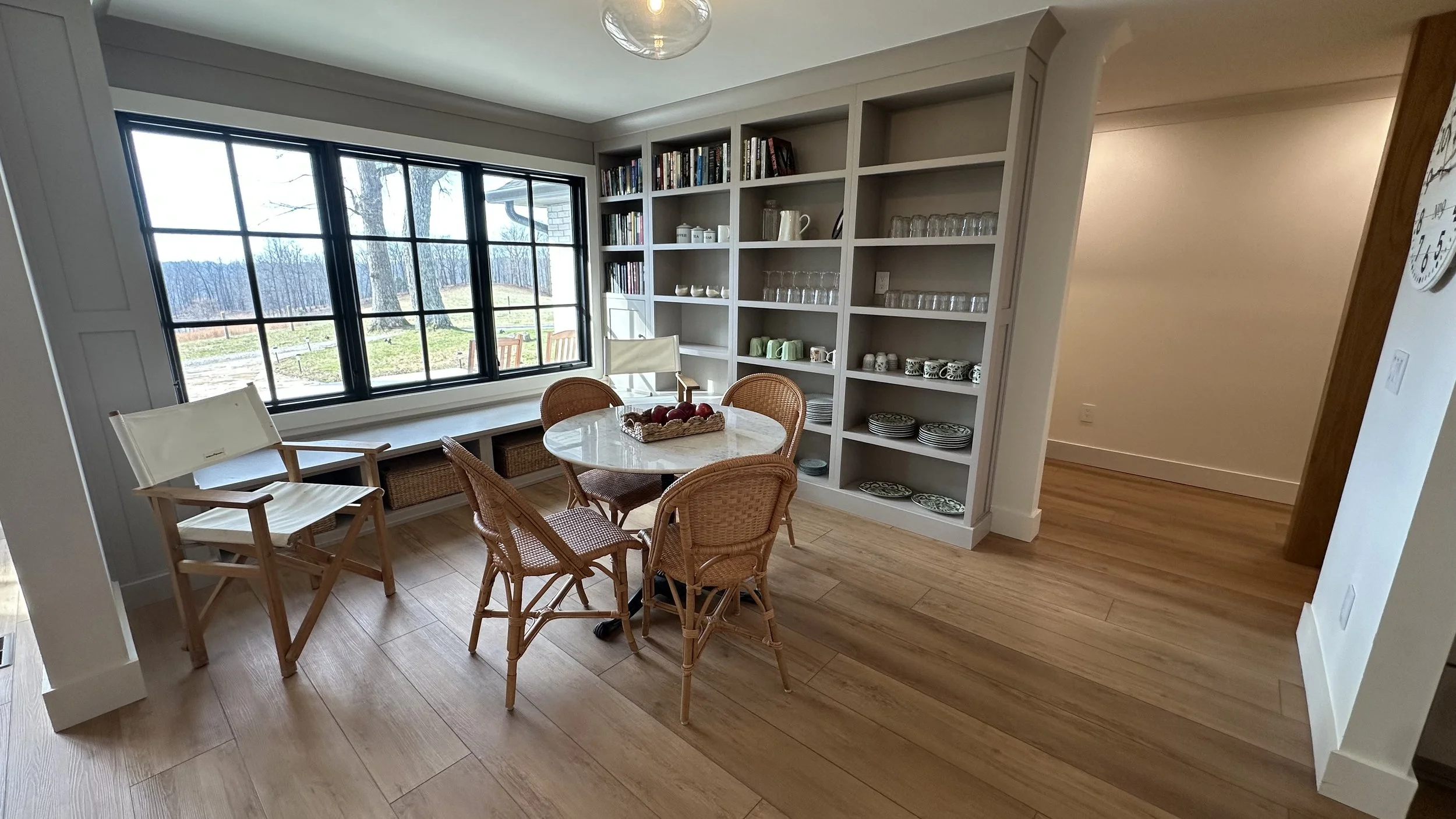 Dining area with a round table, four wicker chairs, a window seat with wicker baskets underneath, large window showing an outdoor view, and a built-in open shelving unit with dishes, glasses, and books.