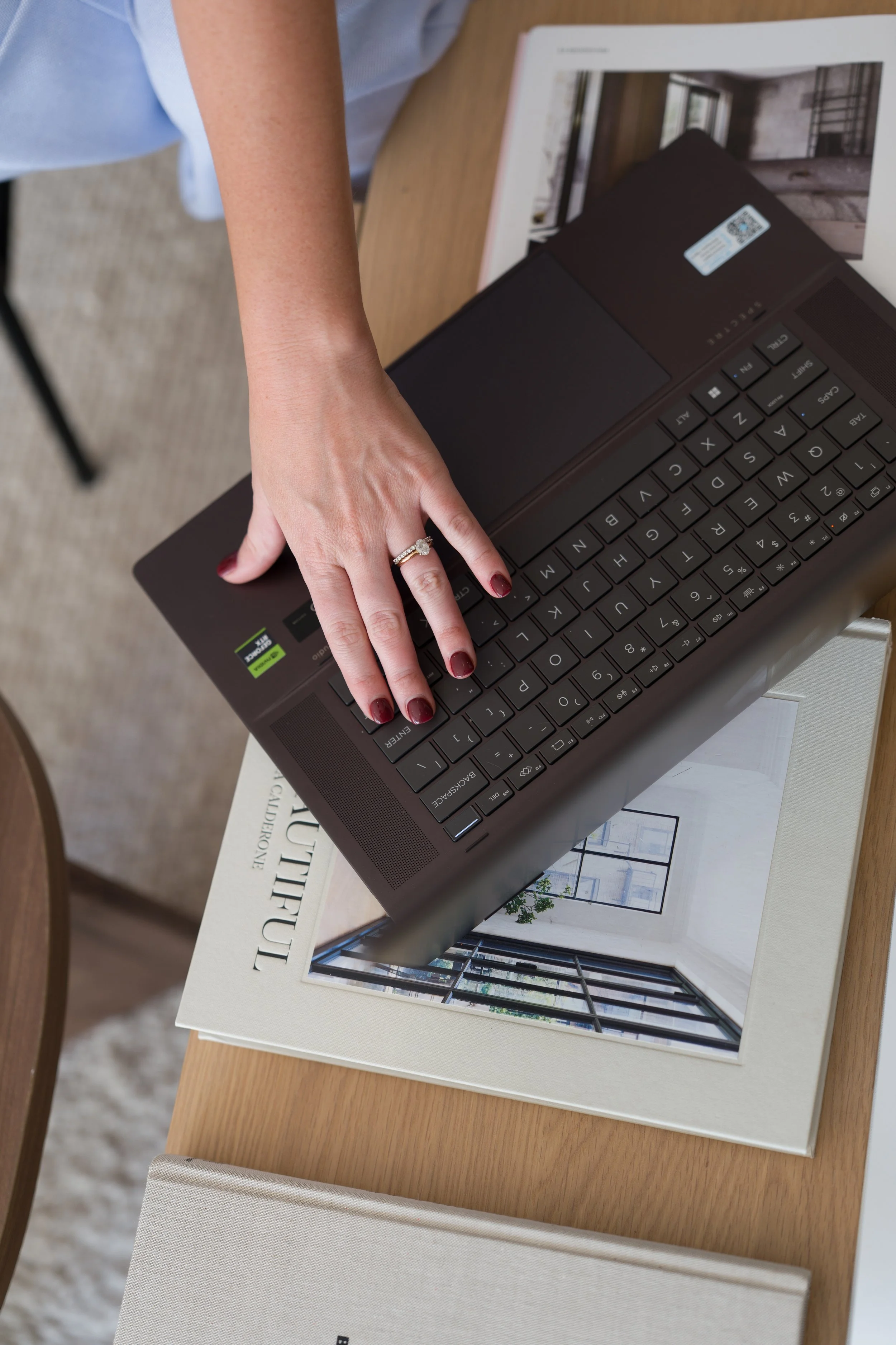 Person sitting on a chair with arms crossed on lap, wearing a black outfit, holding a magazine. The setting appears to be a cozy room with brown chairs and a beige carpet.