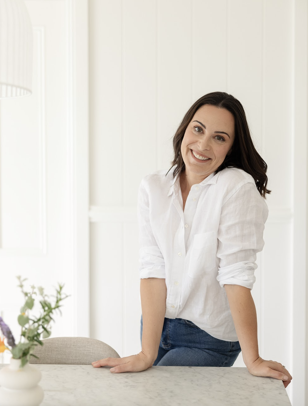 A woman with long wavy auburn hair sitting in a living room, holding a white mug close to her lips, wearing a loose striped shirt and white pants.
