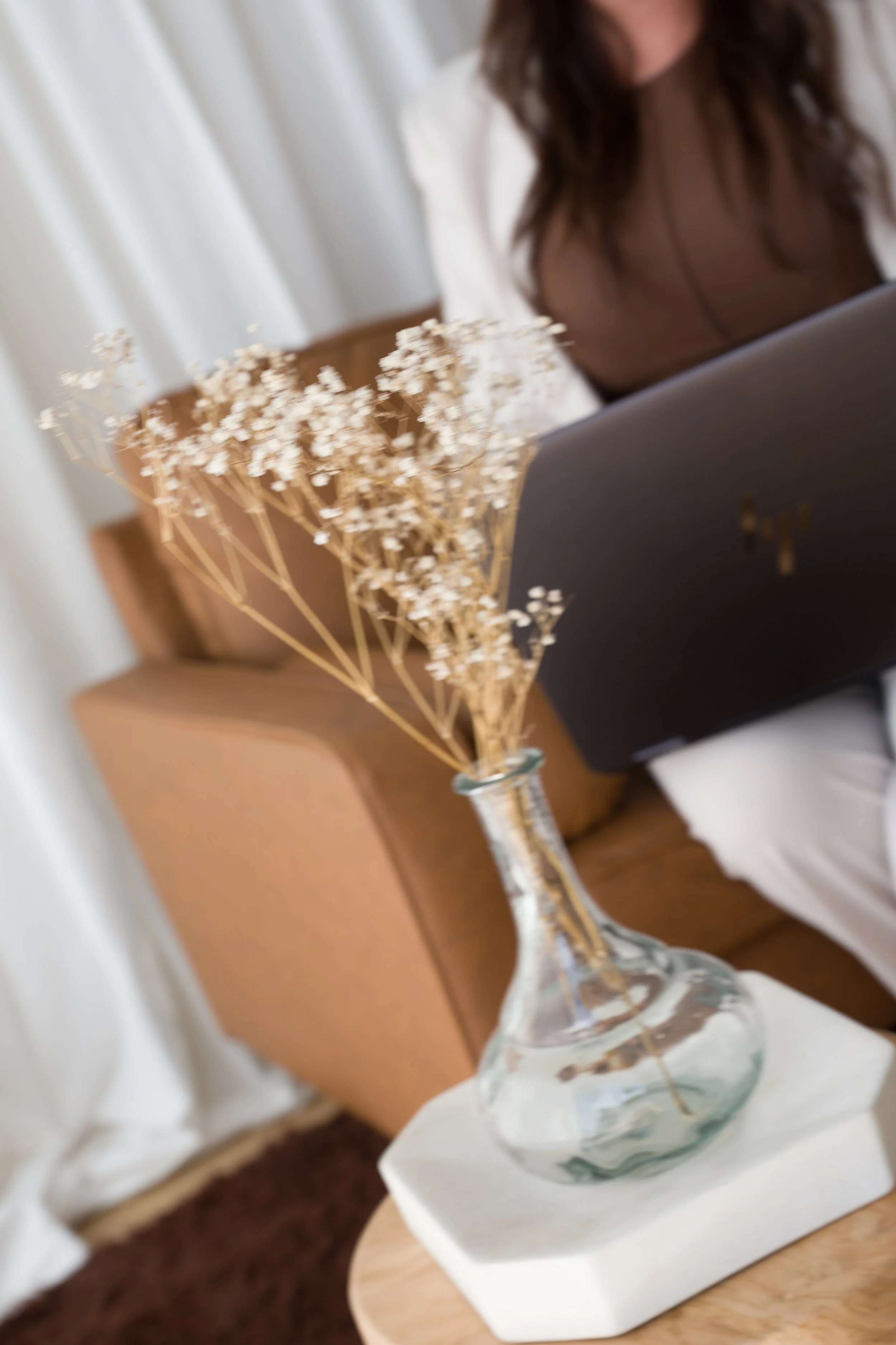 A woman with long, wavy brown hair sitting in a room, holding a HP laptop, wearing a brown top and cream suit