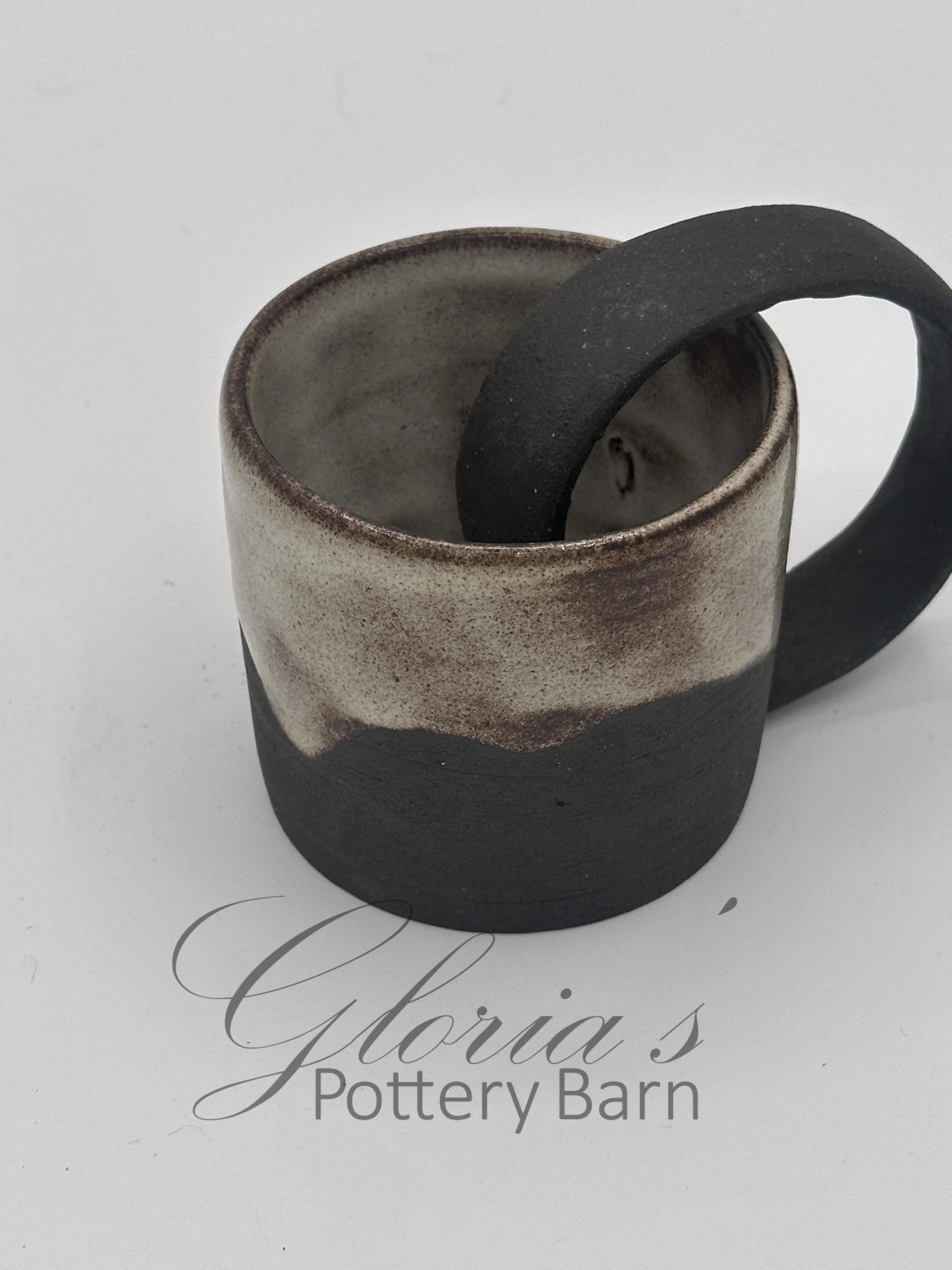 A ceramic mug with a black and beige gradient design and a black handle, placed on a white surface with the text 'Gloria's Pottery Barn' written underneath.