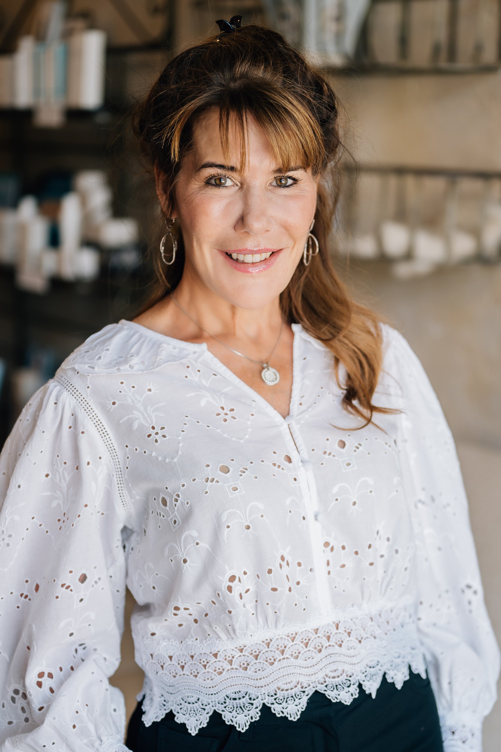 A woman with brown hair and a black hair clip, wearing a white embroidered blouse with lace trim and jewelry, standing indoors with blurred shelves in the background.