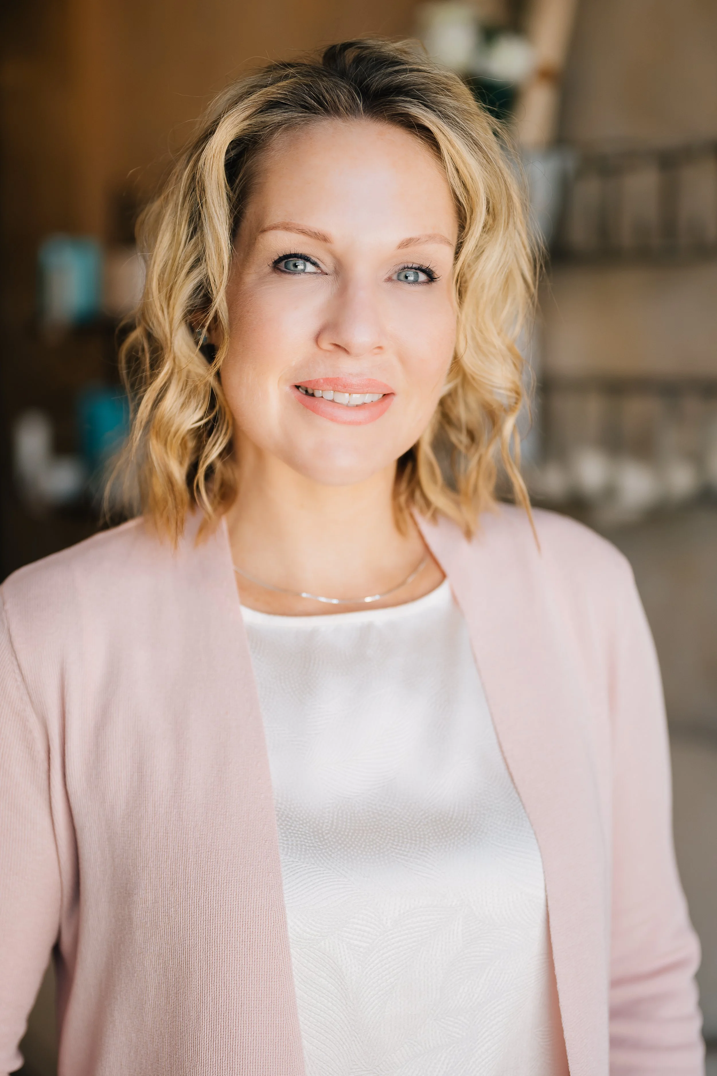 A woman with blonde, wavy hair and blue eyes smiling at the camera, wearing a light pink blazer over a white top, in an indoor setting with blurred background.