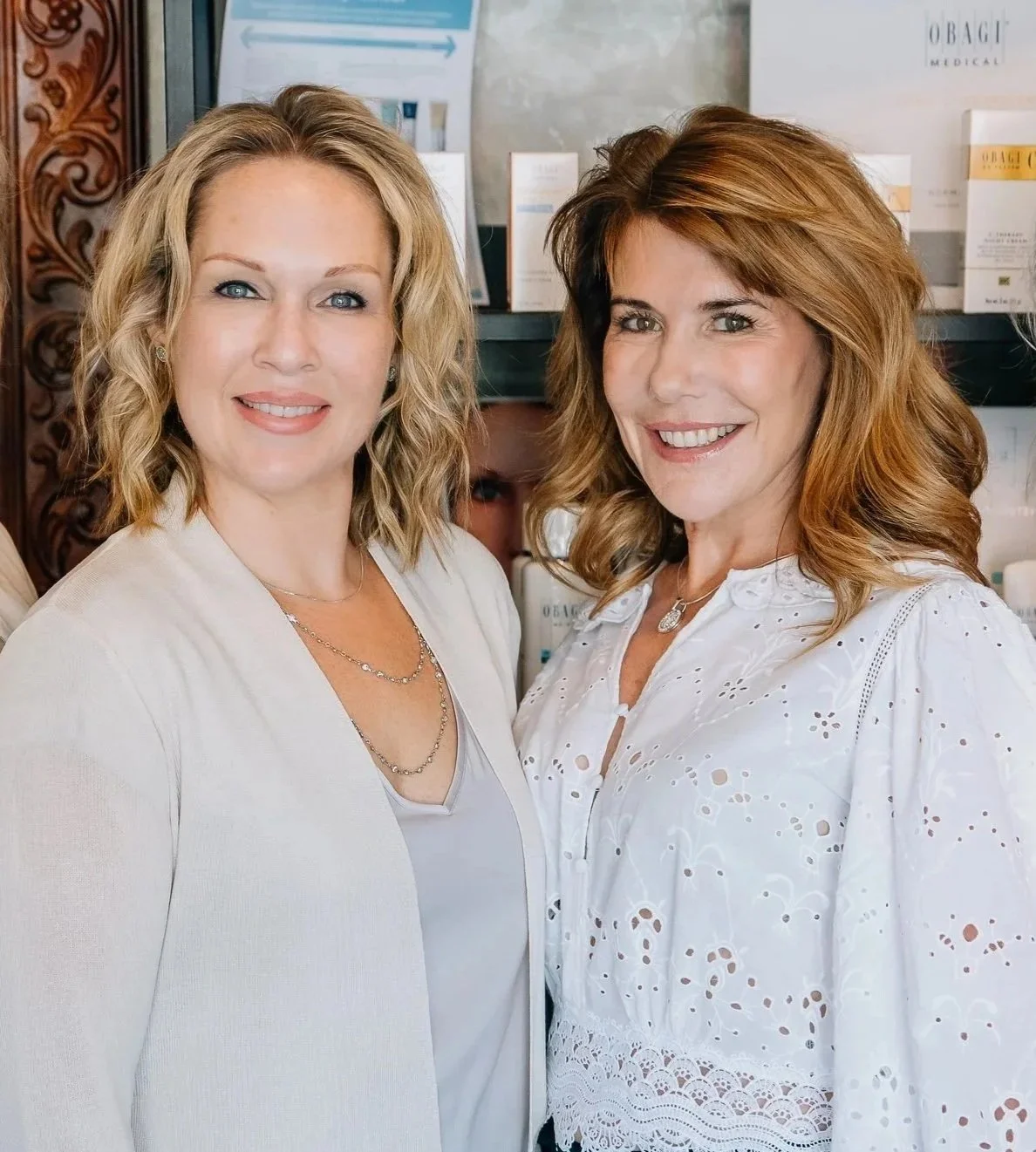 Two women standing together, smiling, in an indoor setting with shelves and skincare products in the background.