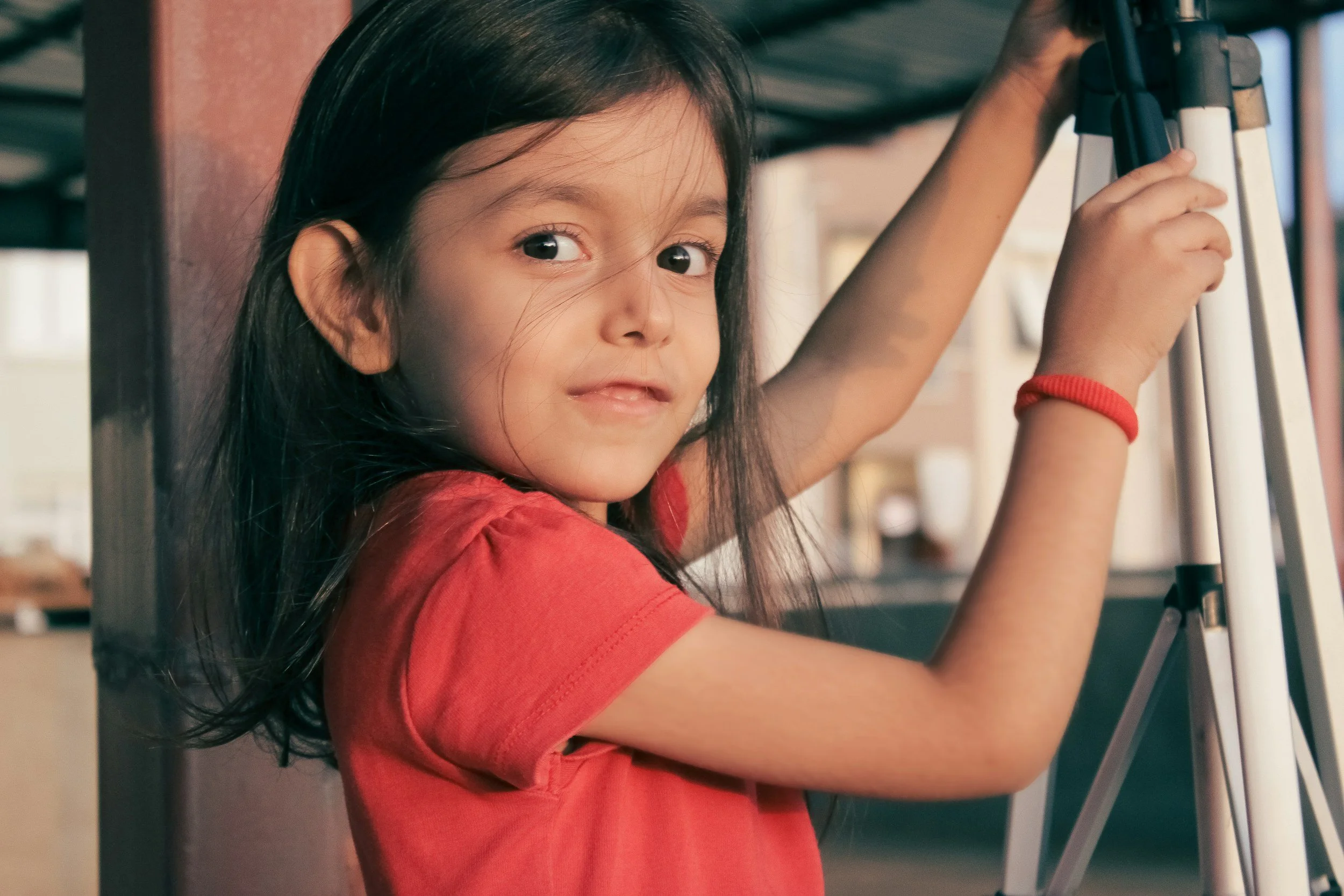 A young girl with long dark hair, wearing a red shirt and a red bracelet, is smiling while holding her hand on a tripod or ladder.
