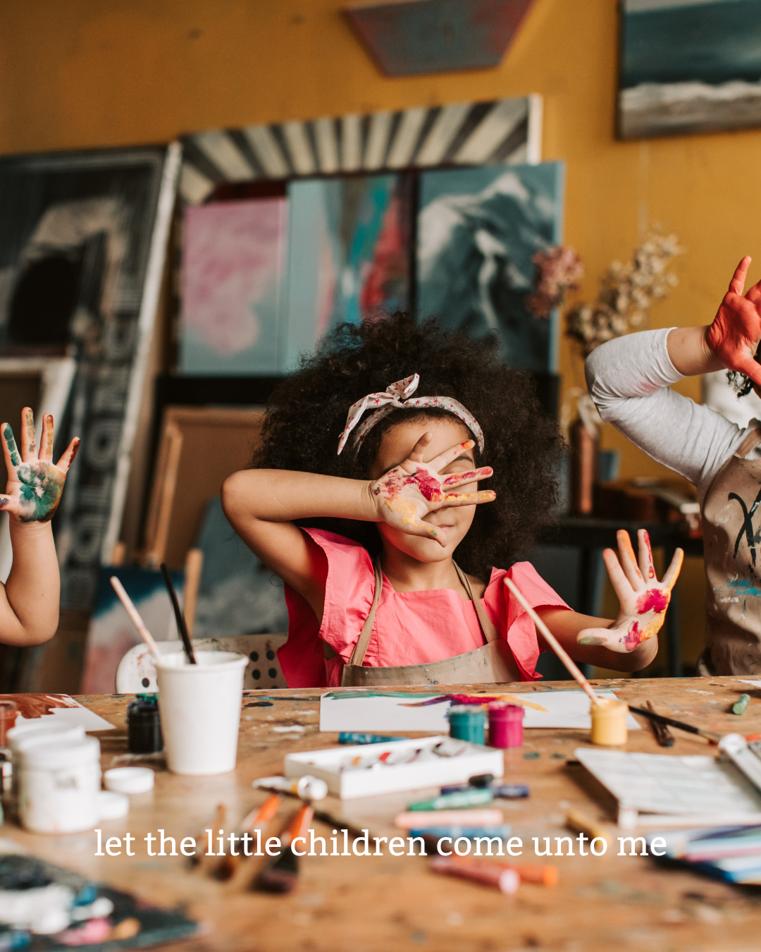 Young girl with curly hair and a headband, wearing a pink shirt, is at a table covered with art supplies, with colorful paint on her hands, covering her face with her hands.