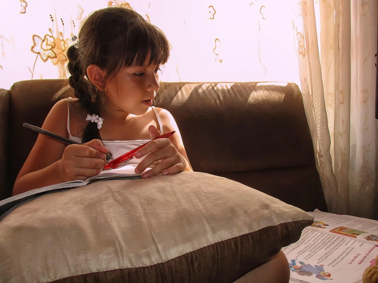 A young girl with dark hair in a braid, sitting on a couch, writing or drawing with a red pen on a notebook resting on a beige cushion. Bright sunlight filters through curtains with floral patterns behind her.