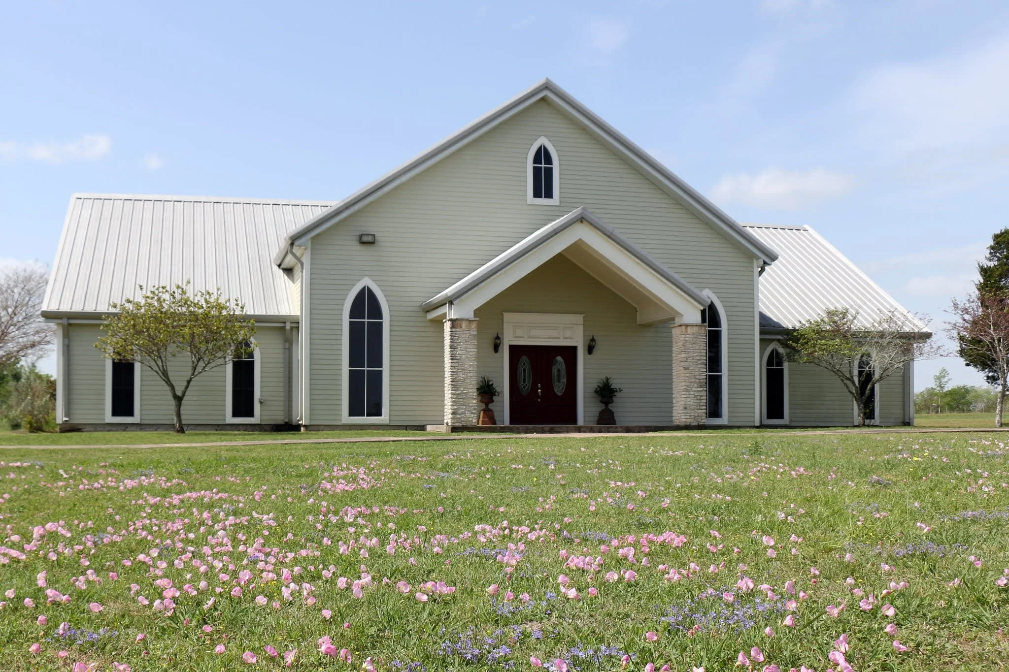 A large, light-colored church building with a metal roof, arched windows, and a front porch with two columns, surrounded by a green lawn with pink and purple flowers and a few trees under a blue sky.