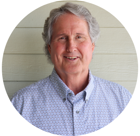 Portrait of a smiling older man with gray hair, wearing a light blue patterned button-up shirt, standing in front of a light-colored wooden panel background.