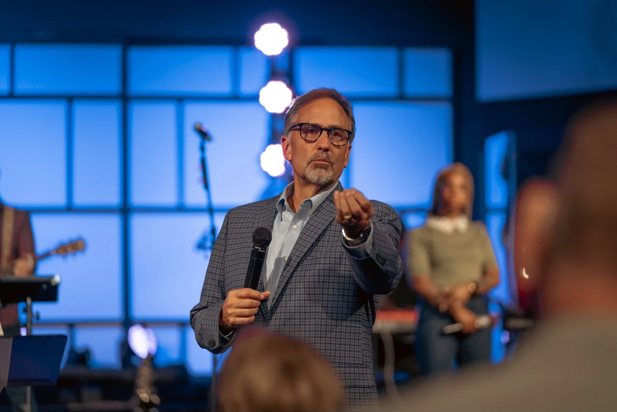 A man with glasses and a beard, wearing a checked blazer, holding a microphone, gesturing with his right hand at a stage with worship leaders behind him and a blue background.