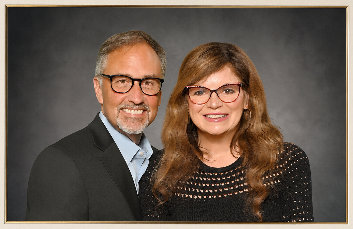 A middle-aged man and woman smiling together, both wearing glasses, against a dark gray background.
