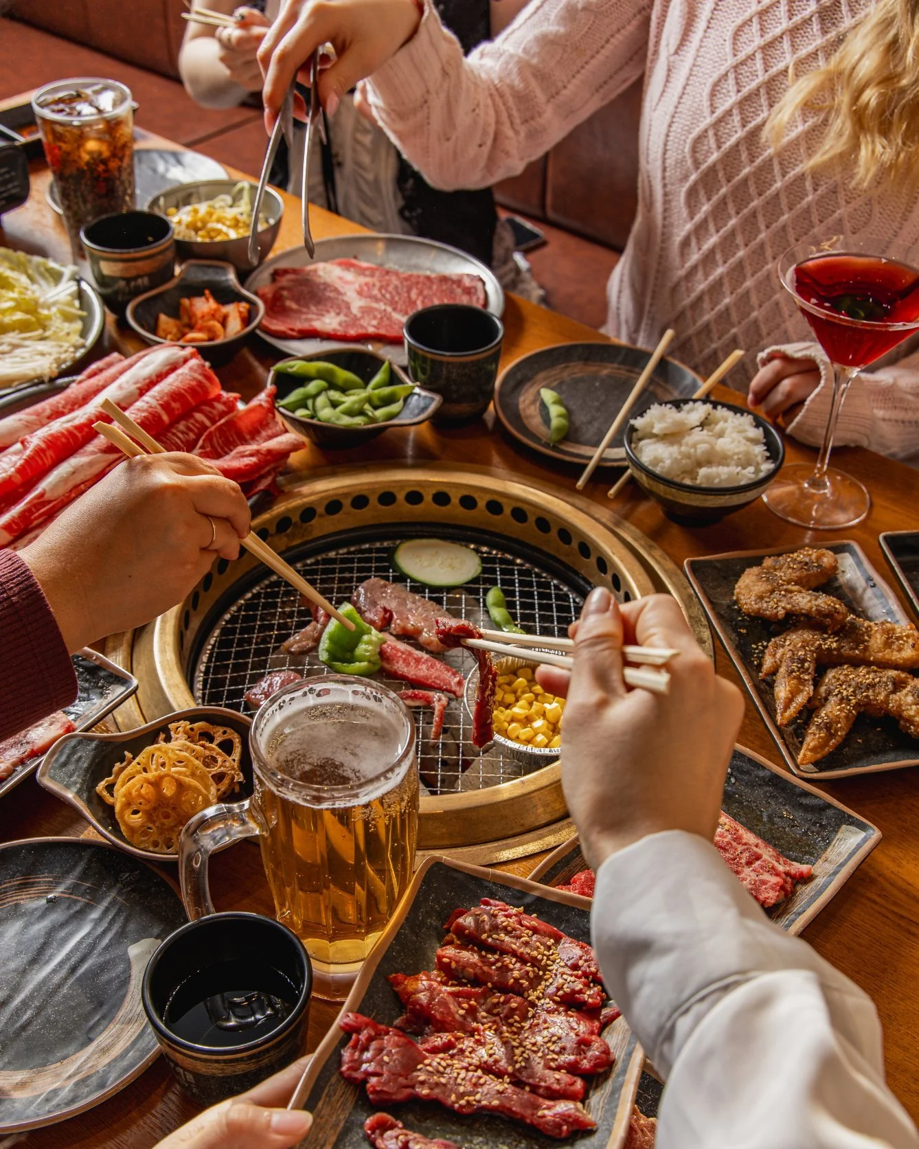 People enjoying a Korean barbecue with various raw meats, vegetables, and side dishes on a table.