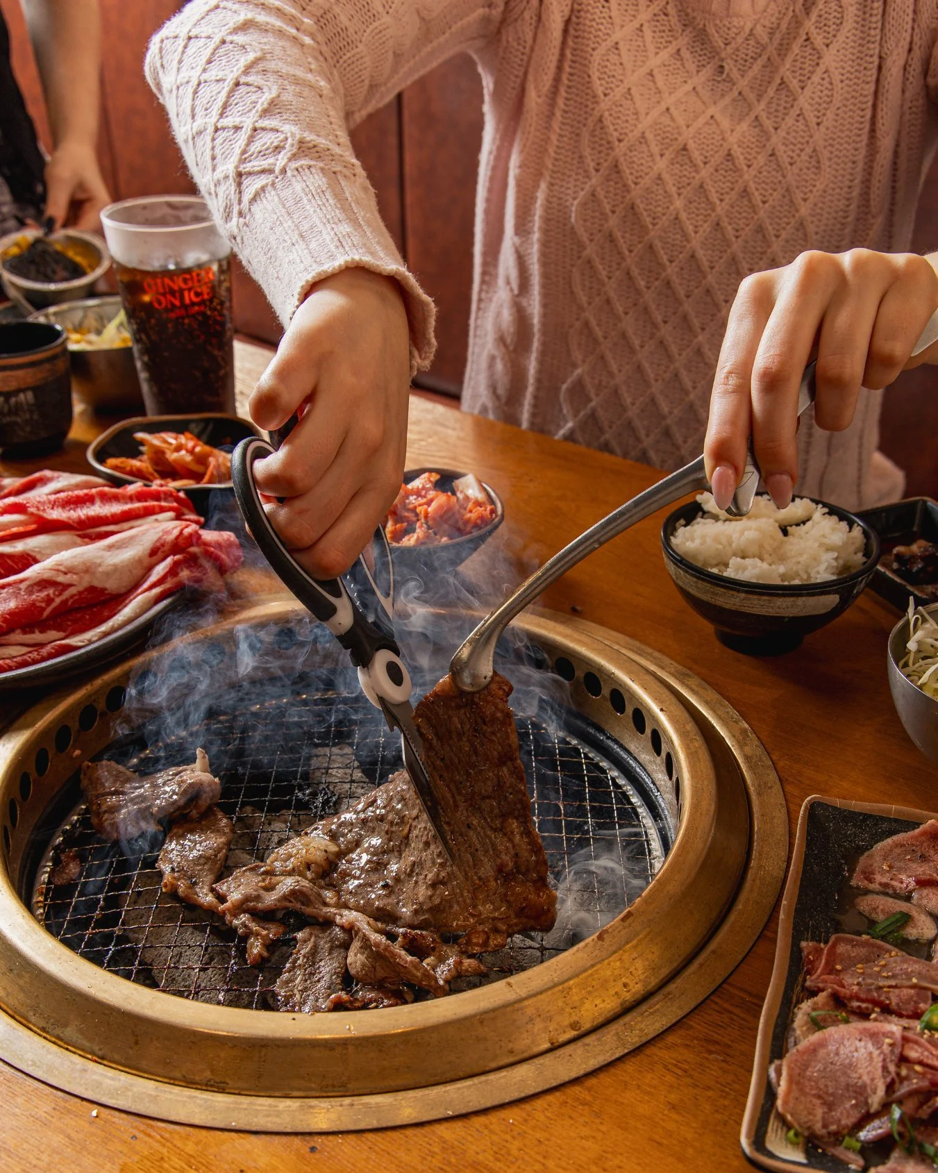 Person grilling meat on a tabletop barbecue, with bowls of rice, sliced meat, and side dishes surrounding the grill.