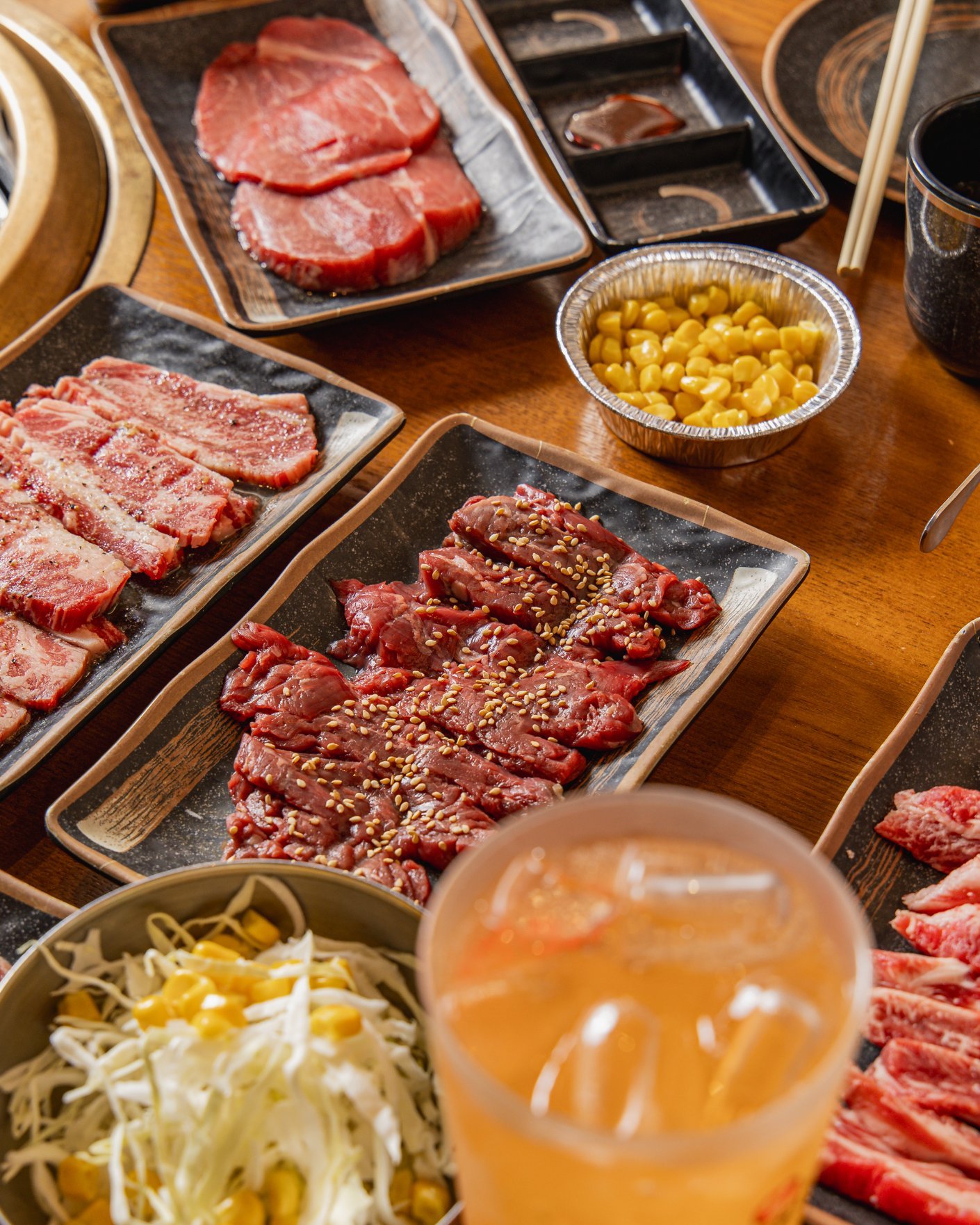 A variety of raw meats, including beef and pork, along with a bowl of corn, shredded cabbage with corn, and a drink with ice on a wooden table, suggesting a meal setting for grilling or barbecuing.