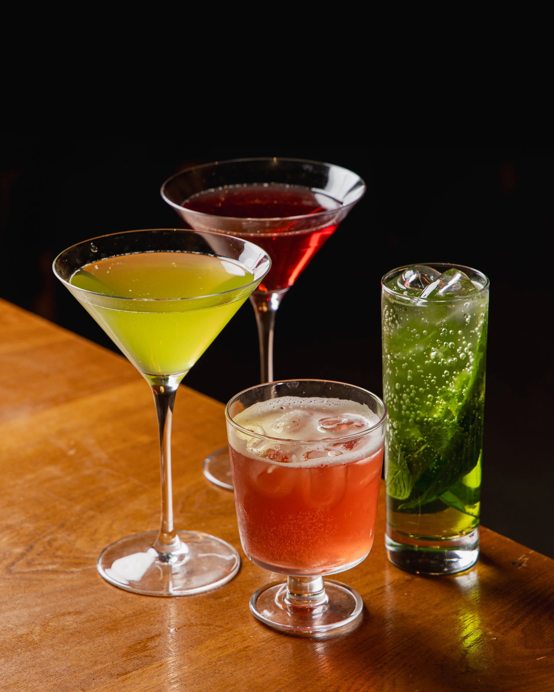 Four colorful cocktails on a wooden bar, with black background. One green in a martini glass, one red in a short glass, one yellow in a martini glass, and one green with ice and mint in a tall glass.
