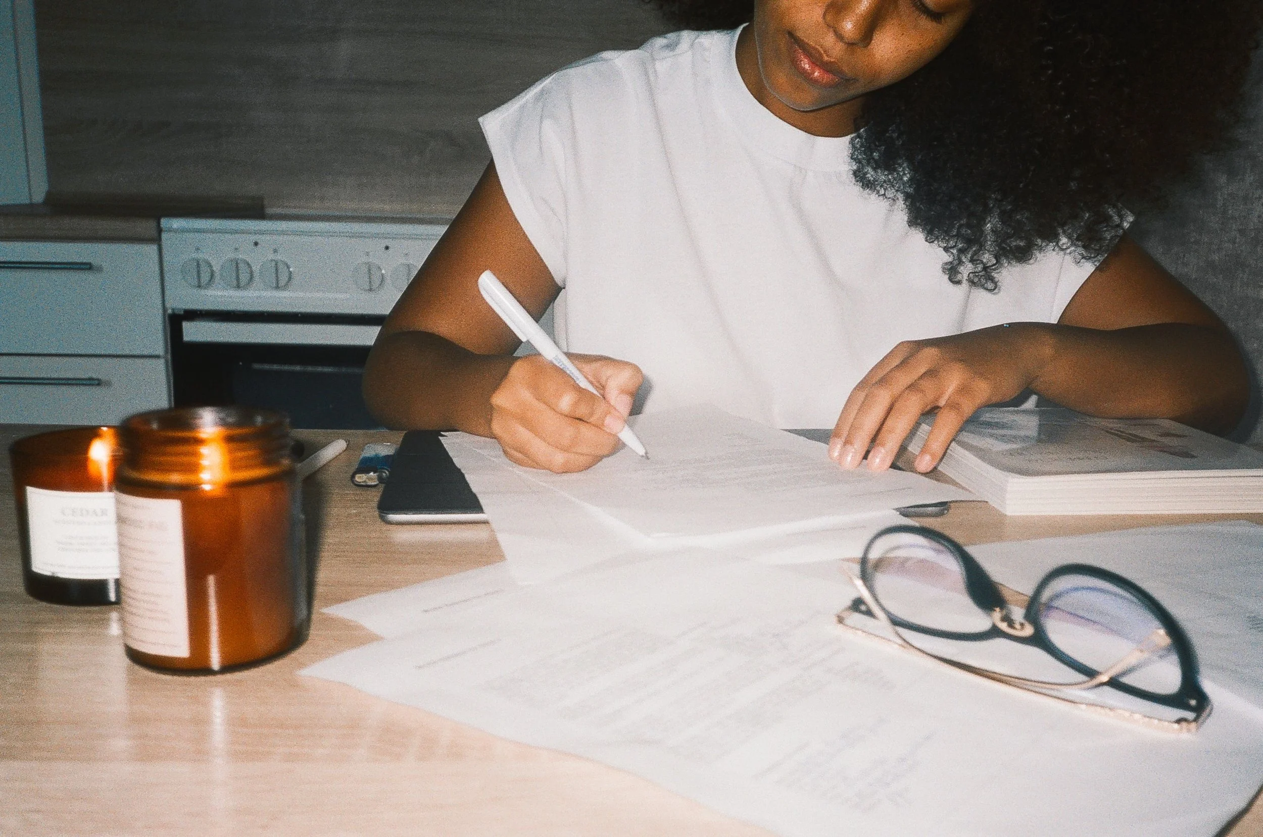 A woman with dark, curly hair sitting at her kitchen table, writing or drawing on paper with a pen. There are black reading glasses, two amber candles burning, and a notebook on the desk.