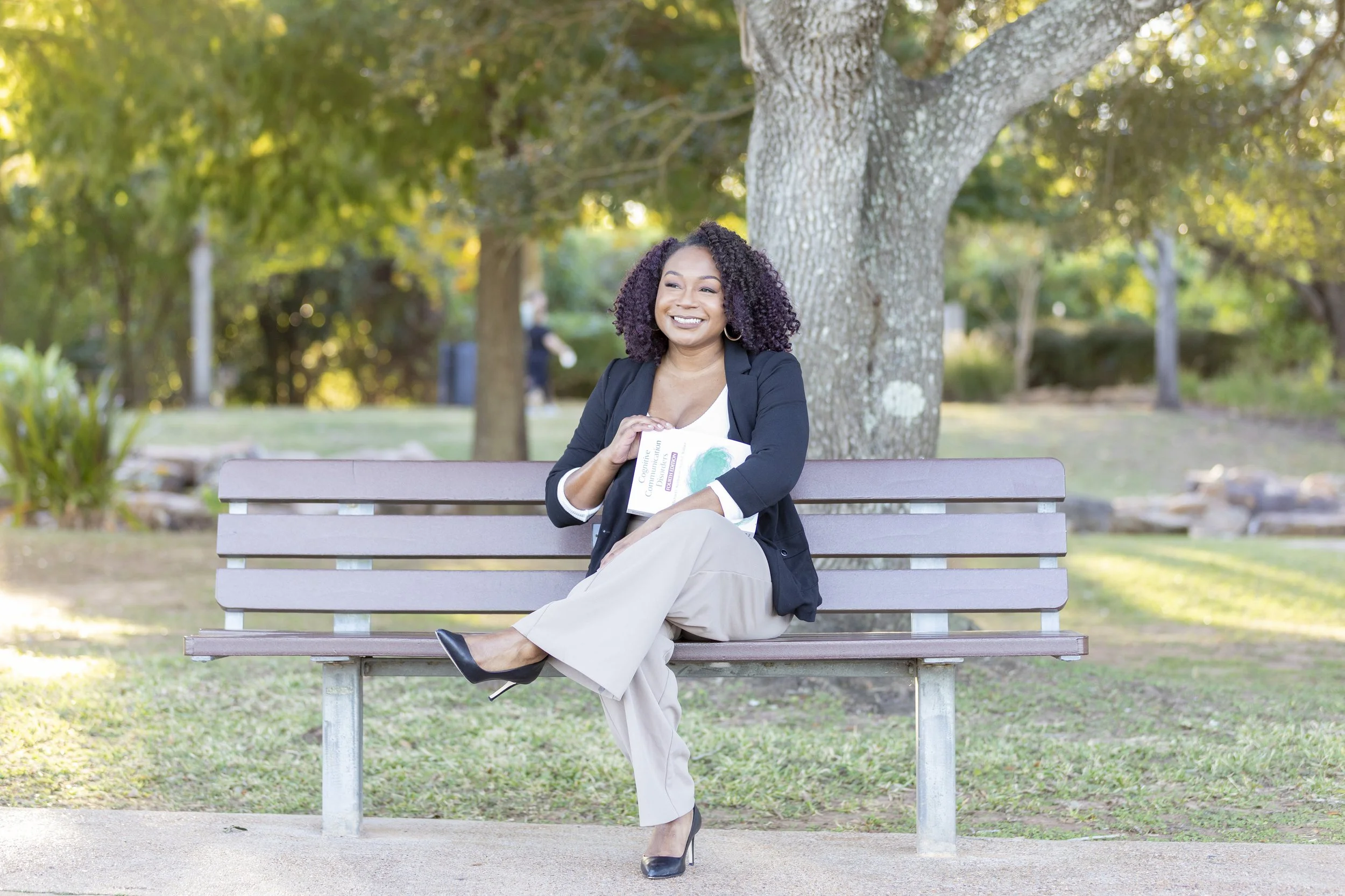 A woman sitting on a park bench under a large tree, holding a book, smiling, wearing a black blazer, beige pants, and black high heels, with green foliage in the background.