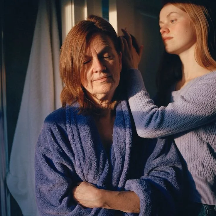 A young woman gently touches the face of an older woman with her eyes closed, both standing next to a window with curtains, in a moment of tender connection.