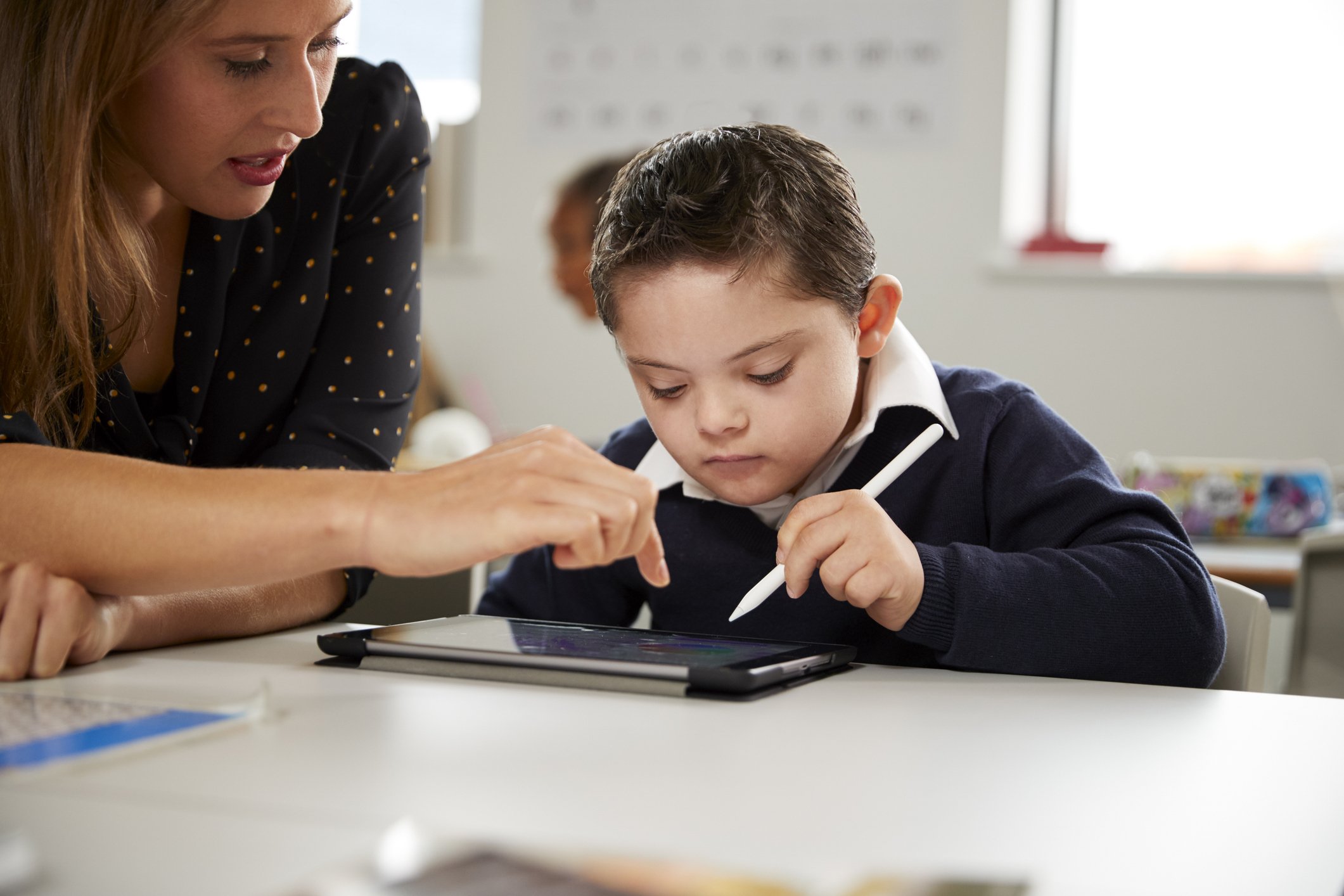 A teacher assisting a young boy using a stylus on a tablet in a classroom.