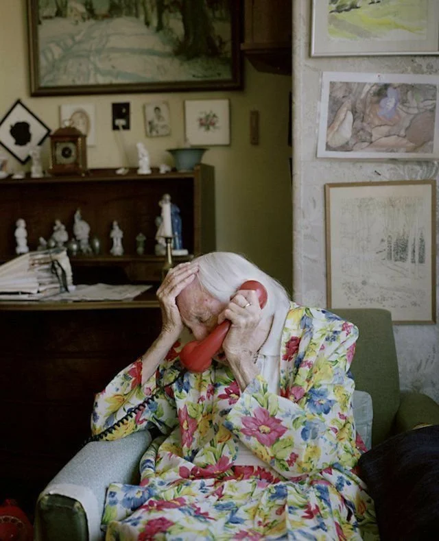 An elderly woman with white hair wearing a colorful floral nightgown, holding a vintage red corded phone to her ear, sitting in a green armchair in a cozy living room decorated with framed pictures and art on the wall.