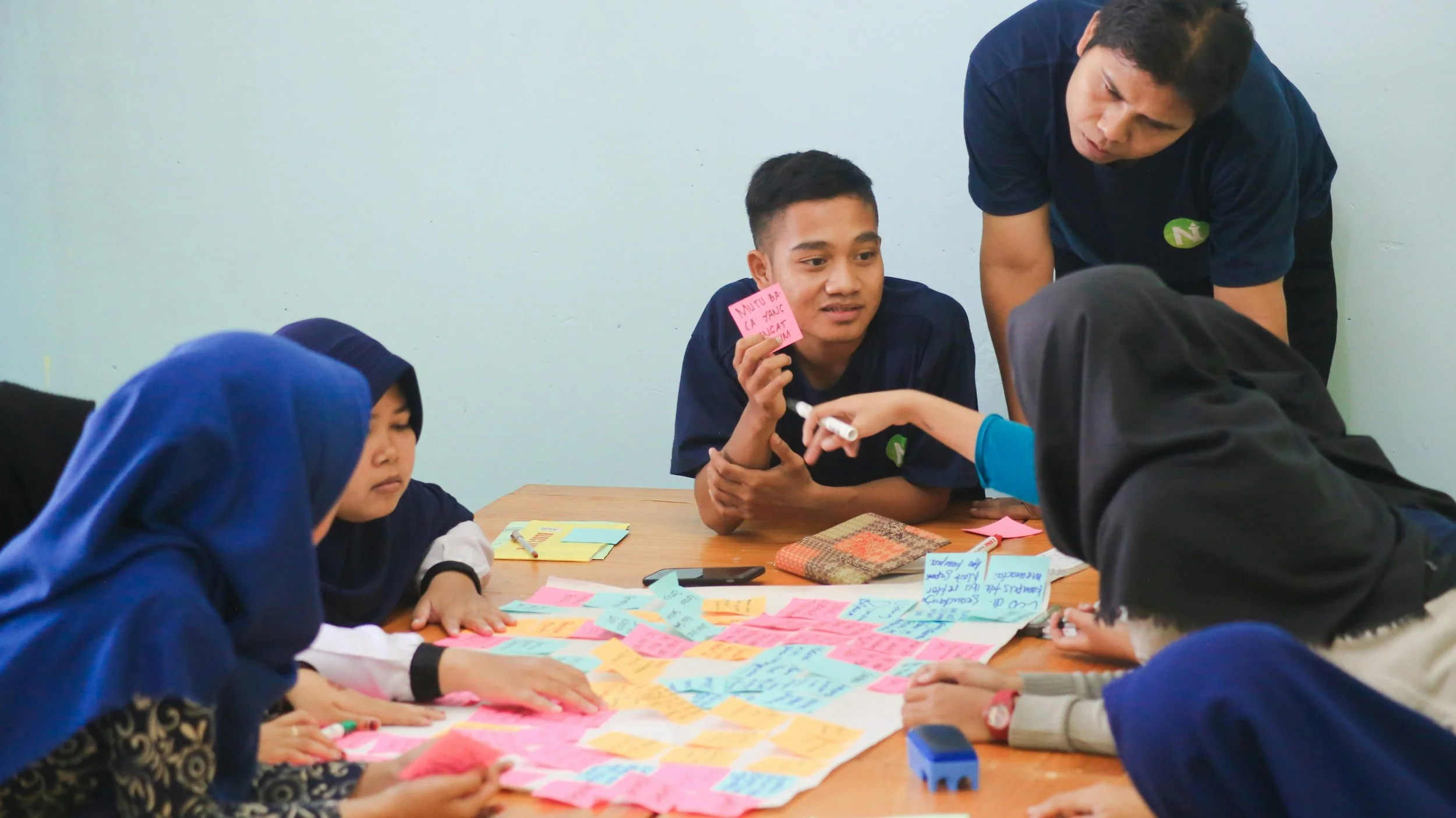 A group of students working on a project with colorful sticky notes on a table, being guided by an instructor in a classroom.