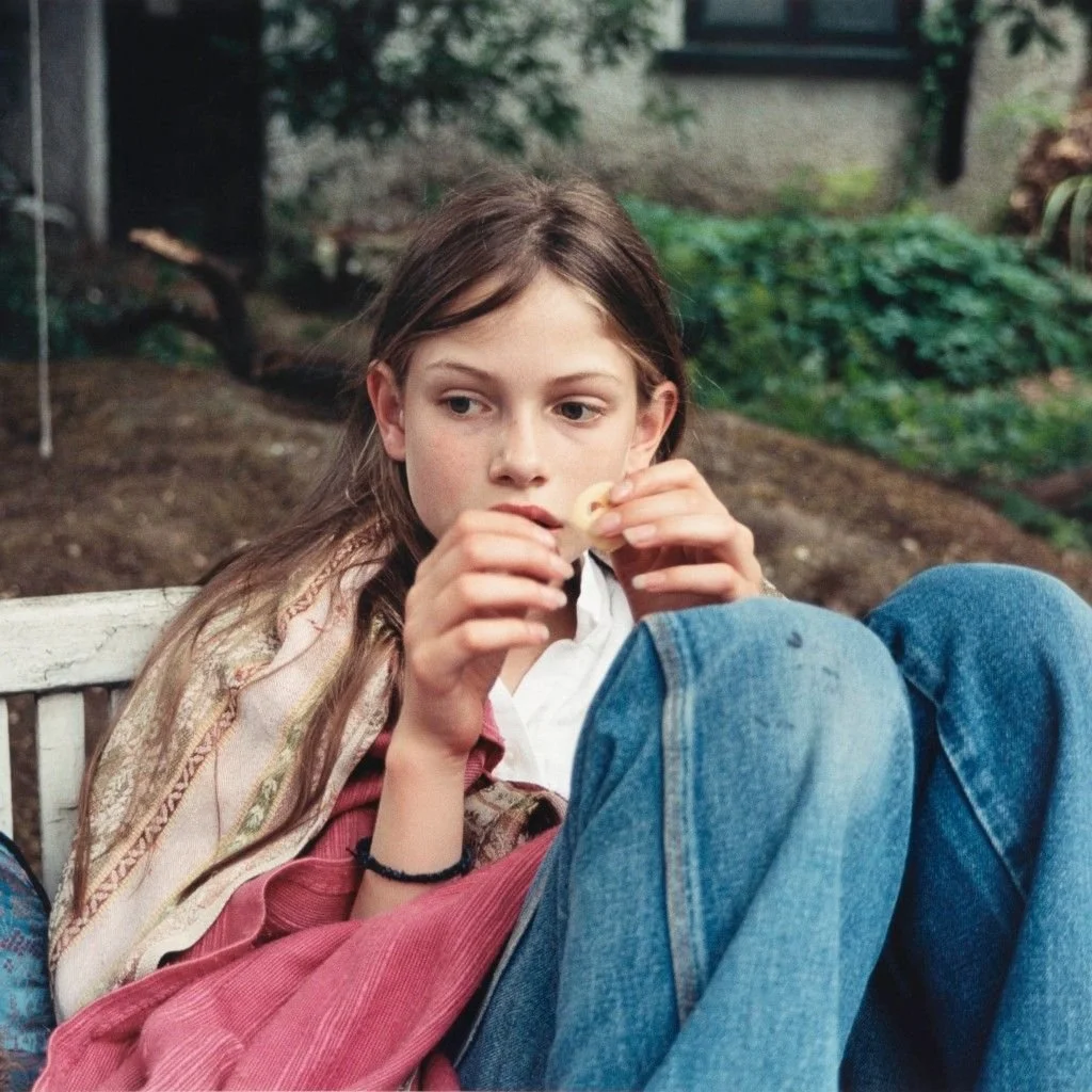 A young girl with long brown hair sitting outdoors on a bench, wearing a white shirt and red pants, holding a potato chip in her hands, with a backdrop of greenery and a building.