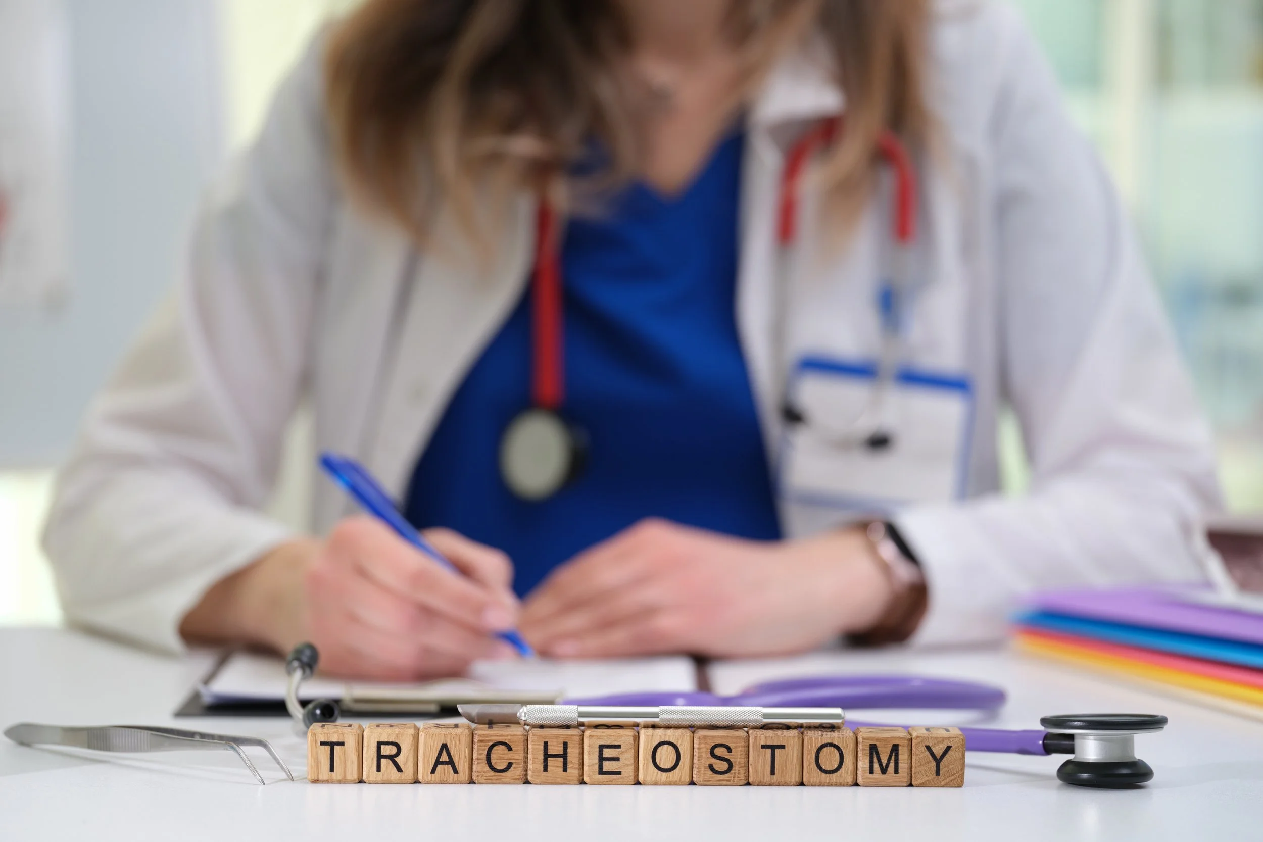 Medical professional writing at a desk with scrabble tiles spelling 'TRACHEOSTOMY', medical tools, and colorful folders.