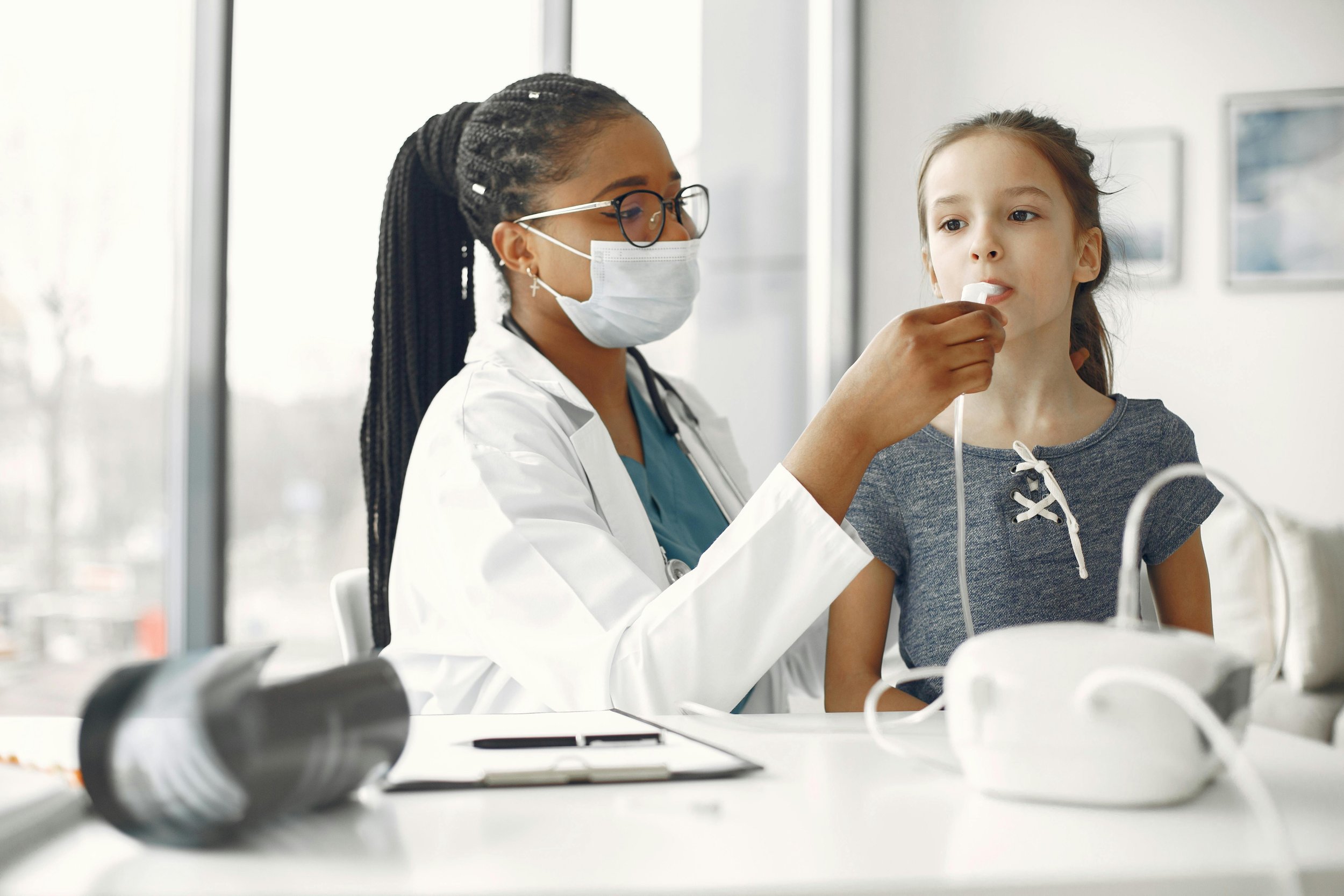 A healthcare professional, wearing glasses, a face mask, and a white coat, is administering a nasal spray to a young girl with brown hair, wearing a gray shirt, inside a clinic with large windows.