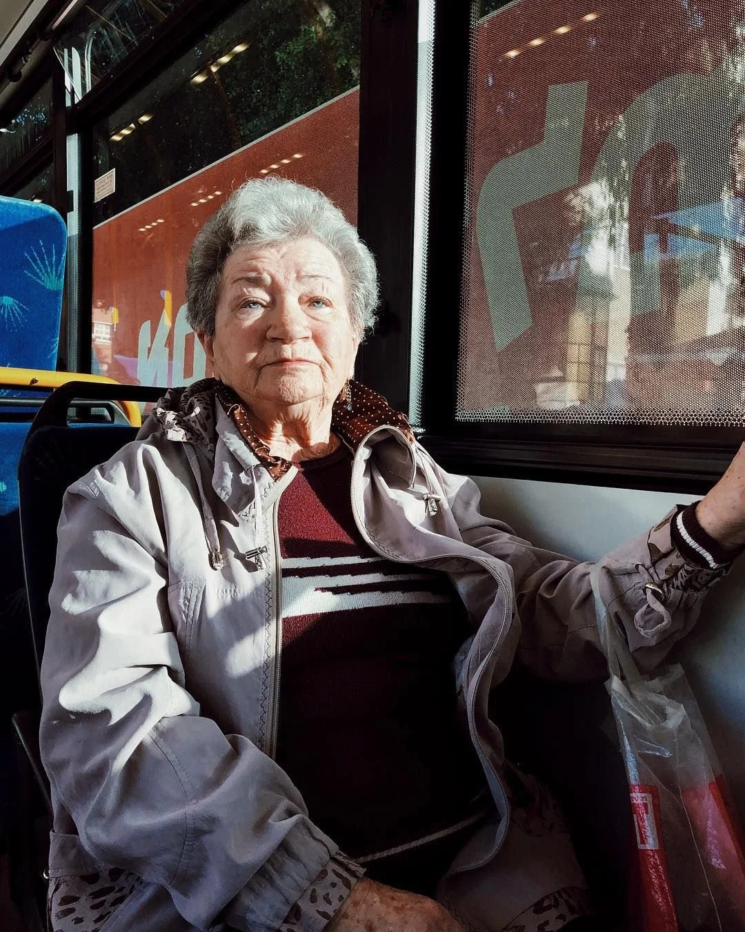 An elderly woman sitting on a bus, gazing out the window with sunlight on her face. She wears a light jacket over a patterned shirt and has gray hair.