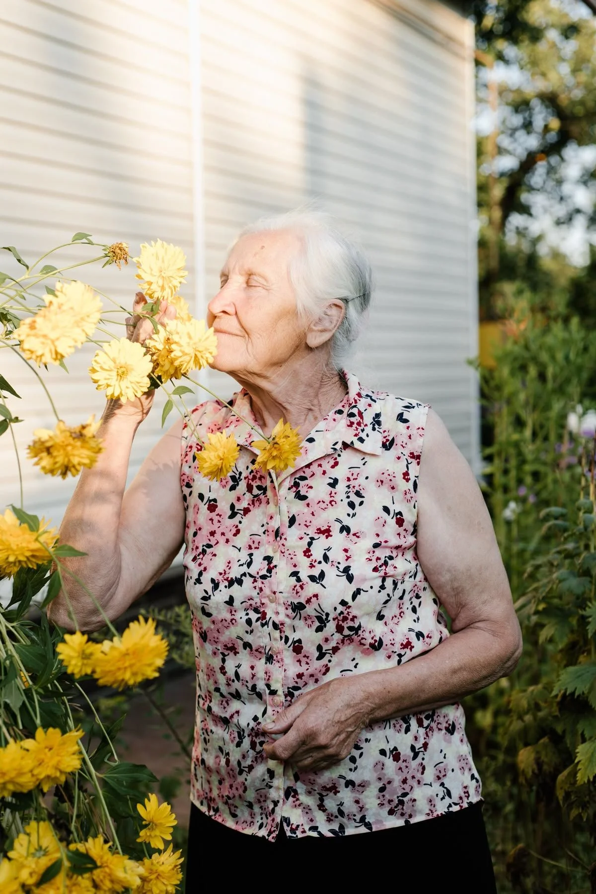 An elderly woman with white hair smelling yellow flowers in a garden near a house.