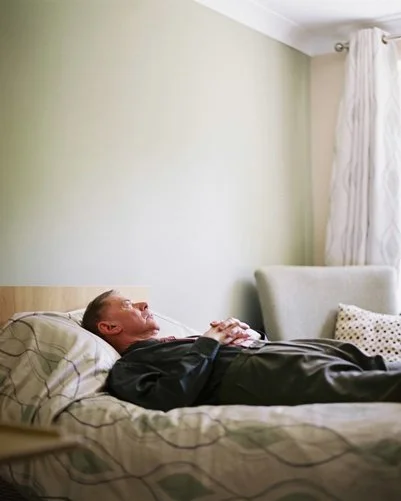 A man lying on a bed with their hands clasped over their chest in a room with a light green wall, a white couch, and a window with cream curtains.