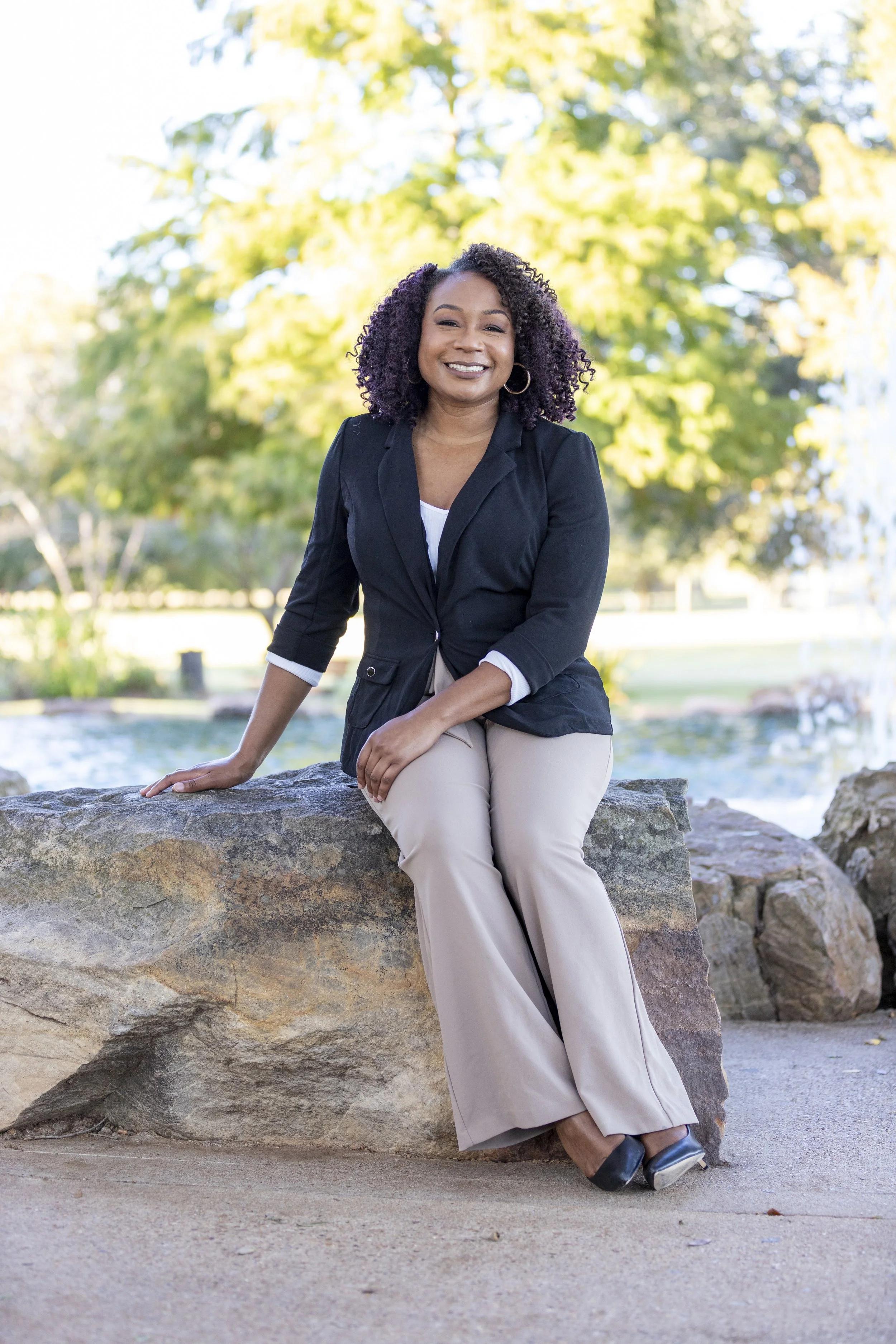 A woman sitting on a large rock outdoors, smiling, with trees and a fountain in the background.