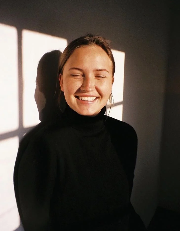 A young woman with long hair smiling with eyes closed, standing in front of a wall with sunlight creating square-shaped shadow patterns.