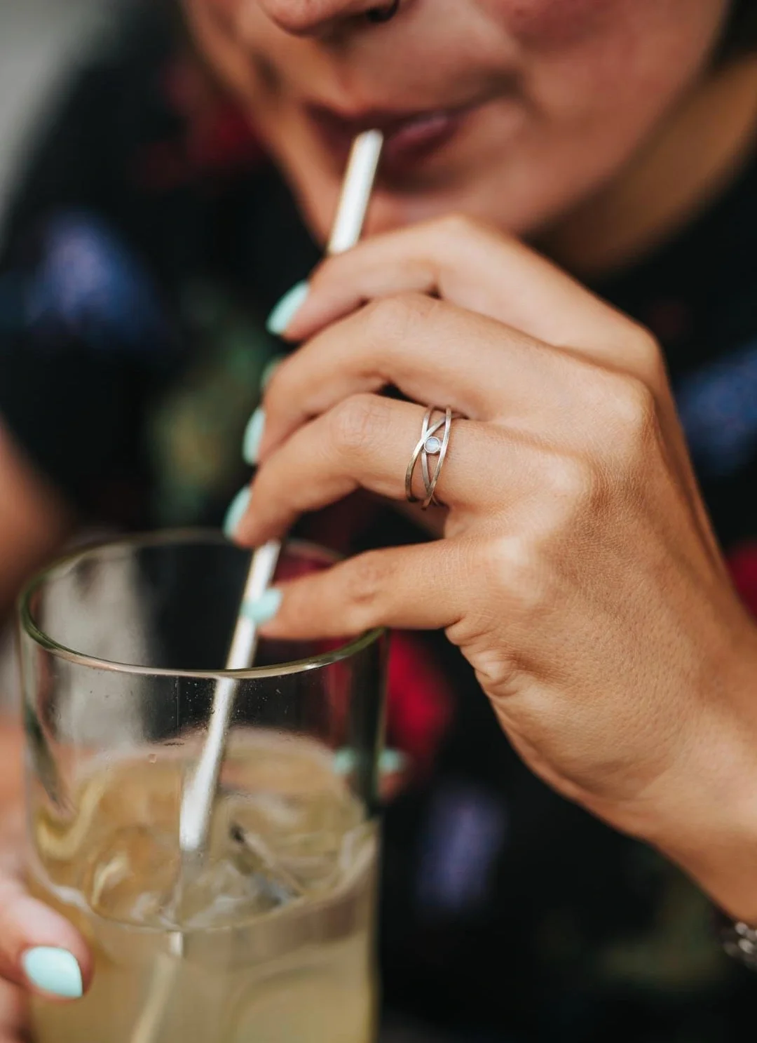 Person sipping a cold drink with a straw, holding the glass with one hand, showing a silver ring with a stone on their middle finger.