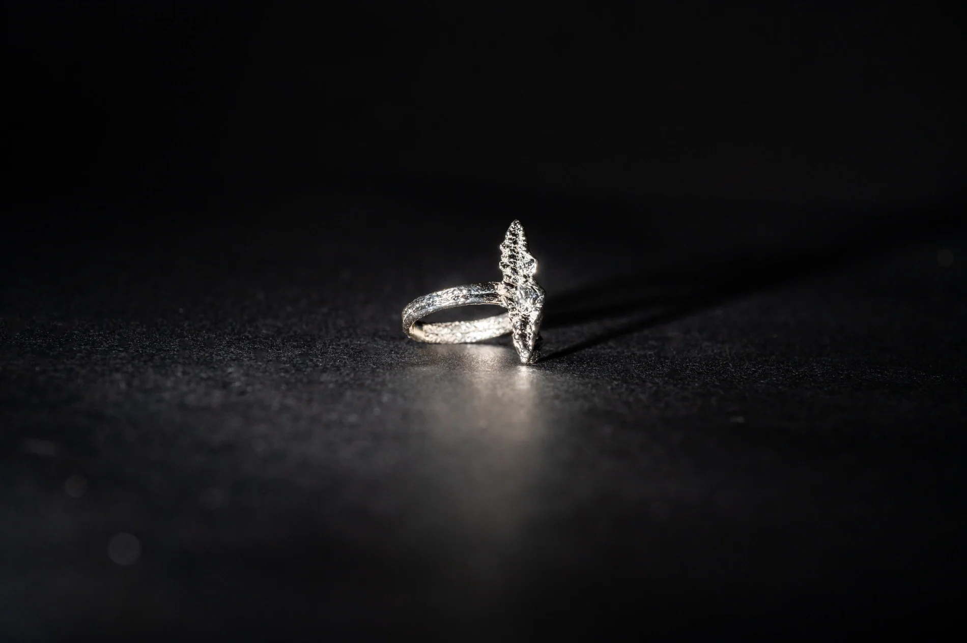 Close-up of a silver ring with a large arrowhead-shaped diamond on a black surface, casting a shadow.