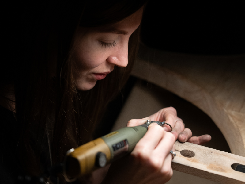 A young woman using a rotary tool to carve or sand wood on a piece of furniture.