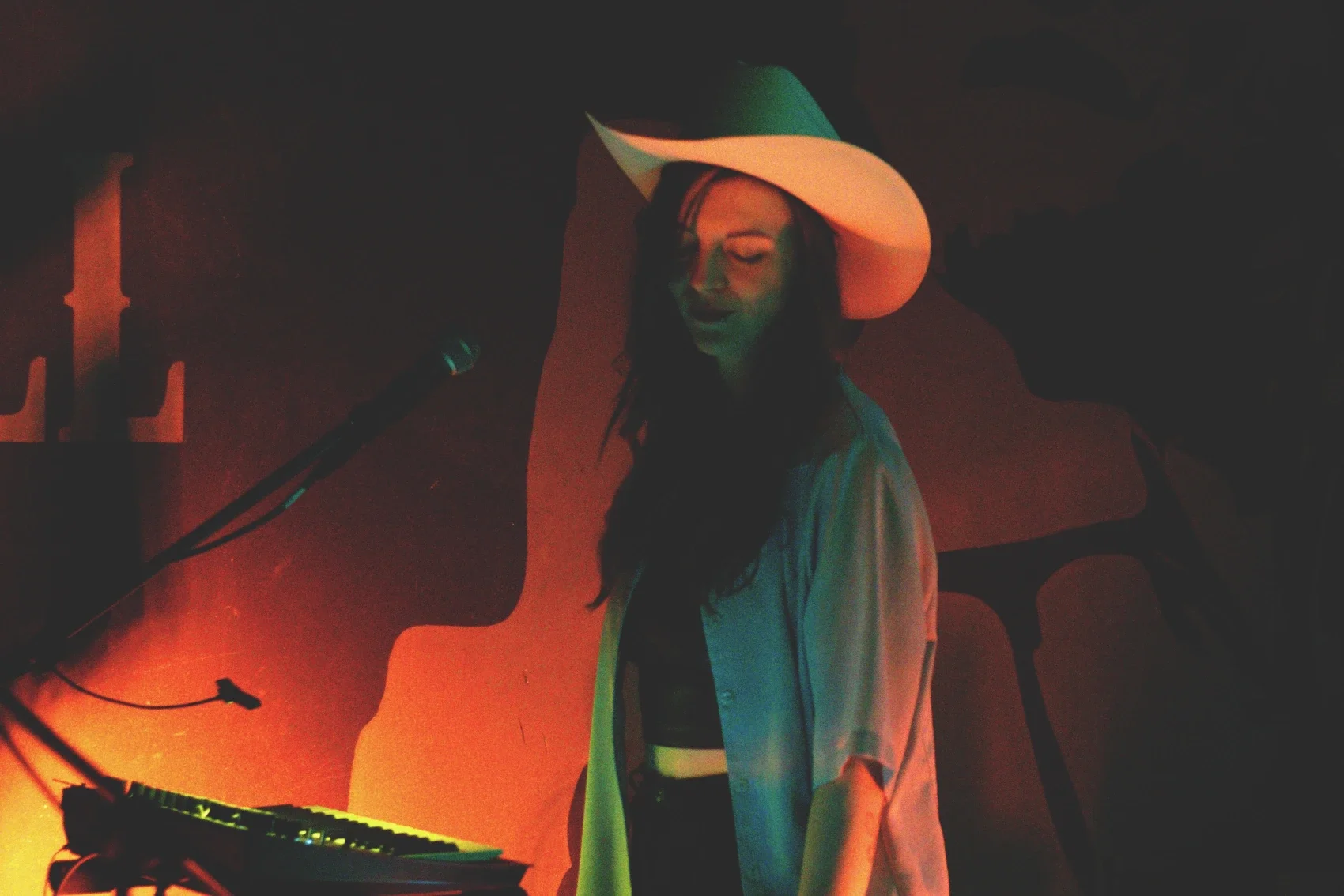 A woman in a large white hat and a light-colored shirt singing or speaking into a microphone on stage, with a keyboard in front of her, clouded in colorful stage lighting.
