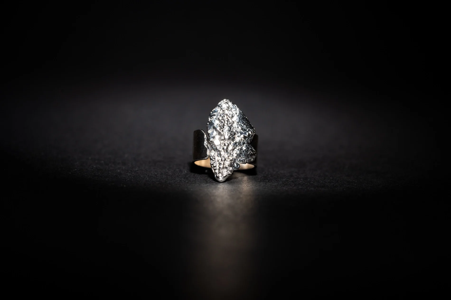 Close-up of a silver ring with a large, jagged, textured surface, illuminated against a black background.